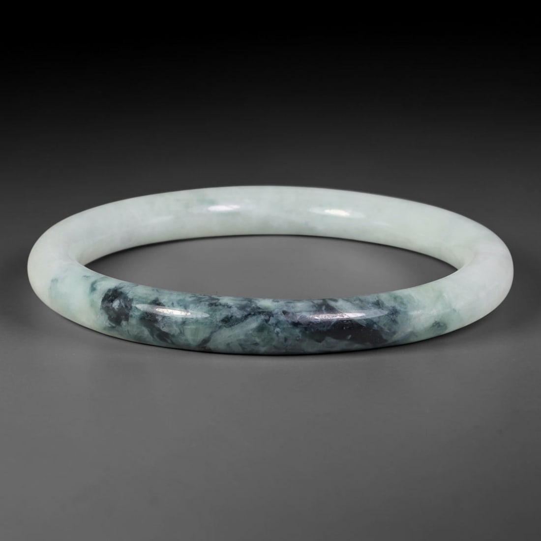 Natural Burmese Type A Jadeite Jade Bangle Bracelet, 13TD: Crafted from natural Burmese type-A jadeite, this slender bangle displays flowing mottled deep green and near-black inclusions set against a pale milky-white base. The smooth rounded hoop form is full