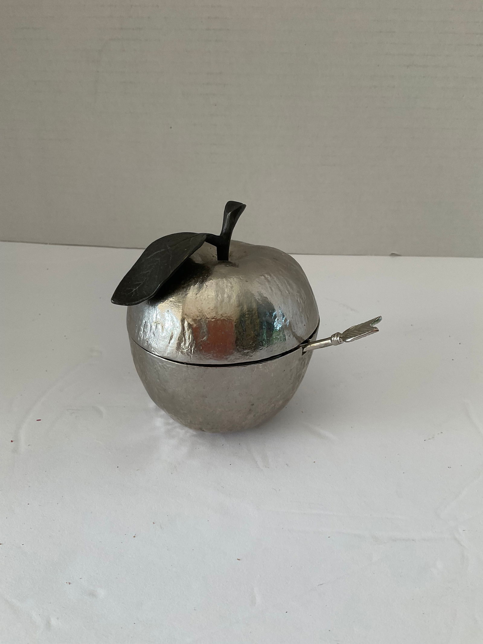 Michael Aram Nickelplated Apple Honey Pot with Red Enamel Interior and an Arrow Spoon: A Michael Aarm Nickelplated honey pot in the shape of an apple. This piece has a natural appeal in the form of an actual apple comple with leaf and stem. The interior is a deep red and the included sp