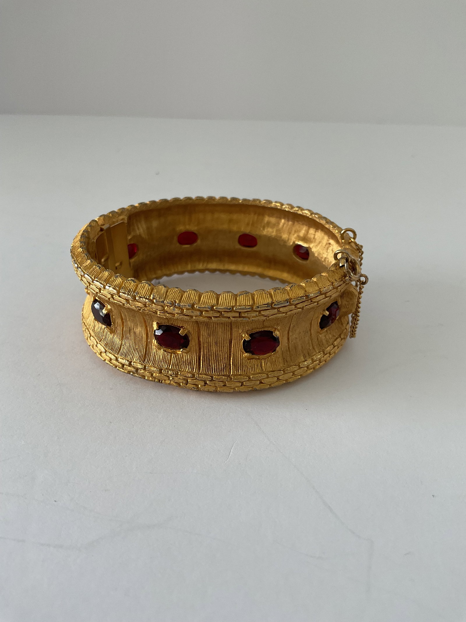 Impressive Hattie Carnigie Signed Hinged Bangle: A very impressive Hattie Carnigie gold tone hinged bangle. It has faceted red gemstones atop a texxtured finish. It measures 3 x 2 x 1" and comes with a safety chain. Minor wear to the finish