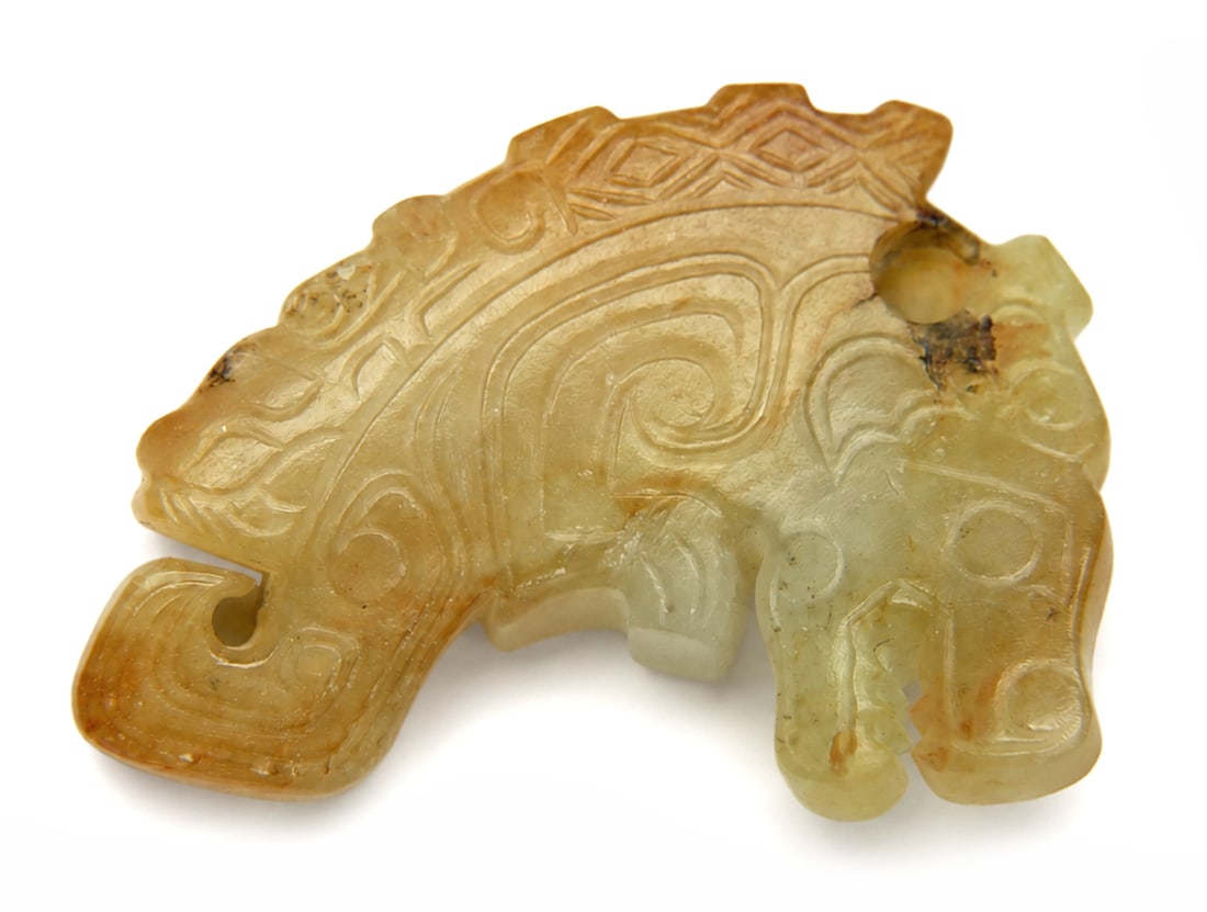 A carved yellowish jade pendant: Dimensions: Length 4.8 cm. Description: A carved yellowish jade pendant in stylised animal form, the surface incised with scrolling decoration and archaistic linear motifs. The warm tone of the stone