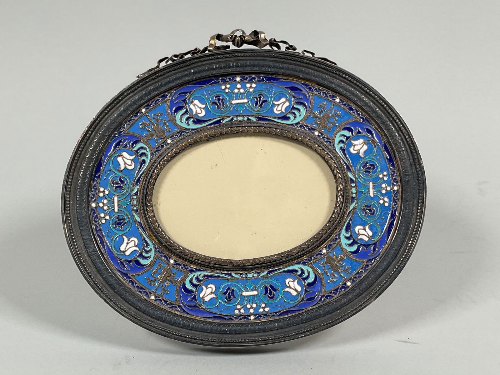 French Champleve Enamel Oval Picture Frame: h: 5 1/2 x w: 6 1/4 inches (overall) h: 2 x w: 3 1/4 inches (opening) CONDITION: the metal ribbon bow is broken and the kickstand is loose with wired repair