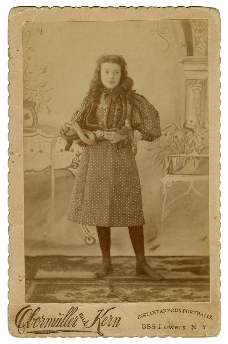 Cabinet Photo of a Child Snake Charmer. New York:: Cabinet Photo of a Child Snake Charmer. New York: Obermüller and Kern, ca. 1900s. Full-length portrait of a girl with snakes draped over her shoulders. Studio mount with scalloped edges. 6 ½