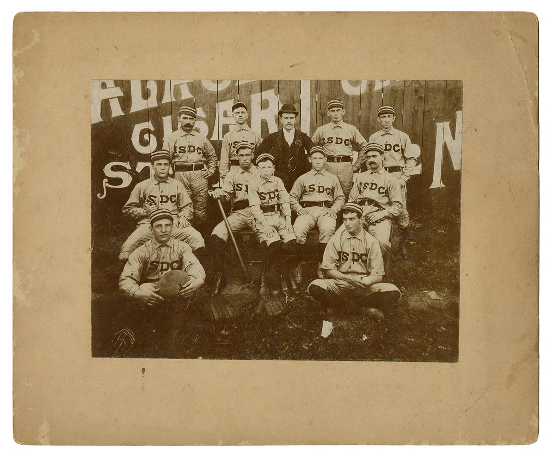[BASEBALL]. Late 19th/early 20th century company team: [BASEBALL]. Late 19th/early 20th century company team photograph. Easton, PA. Sepia-toned photograph depicts a company ball club posed in front of a fence with a painted advertisement. 254 x 356 mm. C
