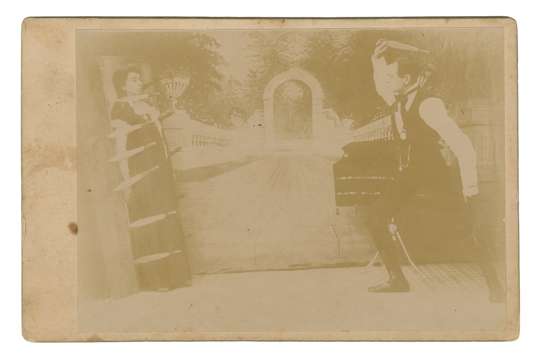 [KNIFE-THROWER]. Cabinet card. N.p., ca. 1880s-90s. Photogr...: [KNIFE-THROWER]. Cabinet card. N.p., ca. 1880s-90s. Photograph on card mount of a knife thrower and his assistant, who stands against a target board with knives outlining her body. 6 ½ x 4 1/8