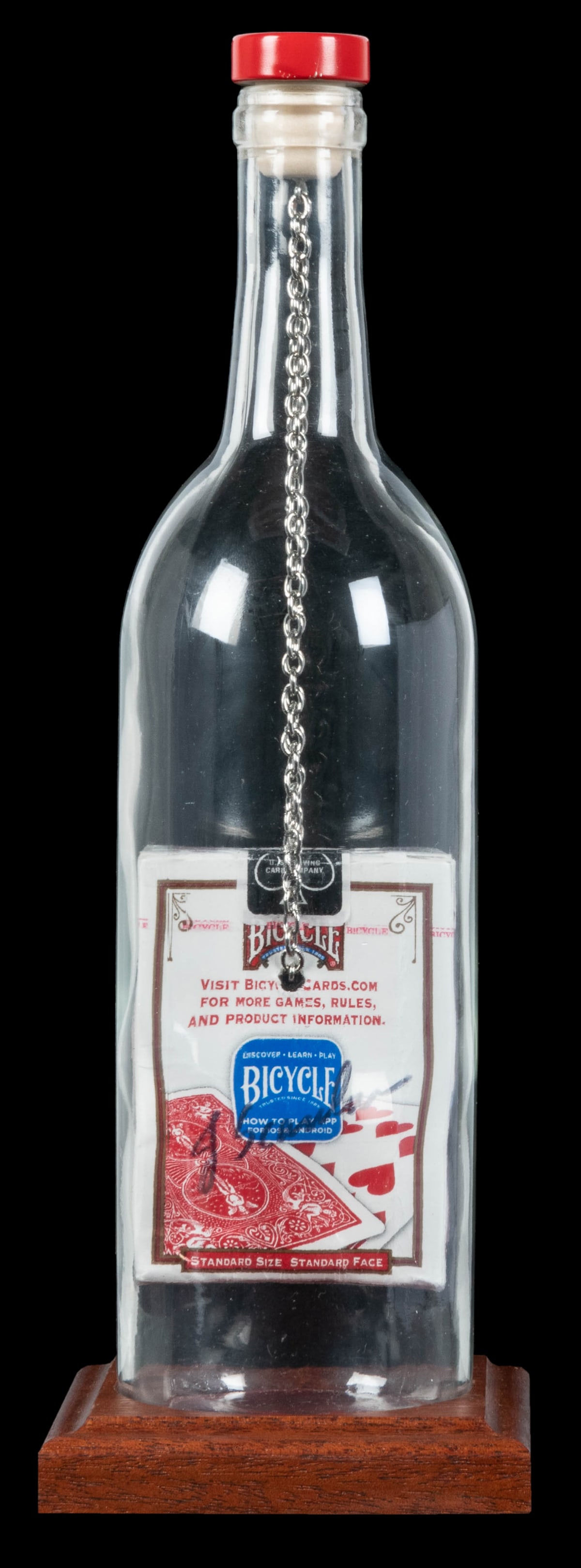 Red Deck Merlot Chained Impossible Bottle.: Red Deck Merlot Chained Impossible Bottle. Arizona: Jeff Scanlan, 2020s. Empty glass wine bottle contains a full sealed pack of cards, threaded on a length of chain running through a hole in the