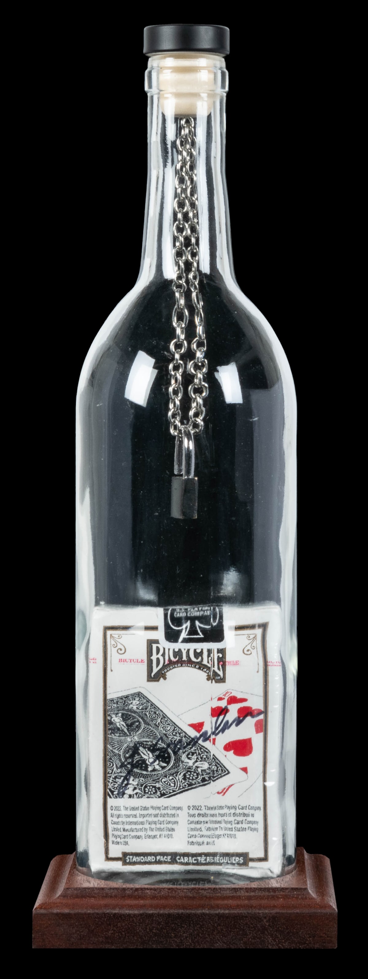 Black Deck Merlot With Lock Impossible Bottle. : Black Deck Merlot With Lock Impossible Bottle. Arizona: Jeff Scanlan, 2020s. Empty glass wine bottle contains a full sealed pack of black Bicycle cards. A chain suspended from the cork inside the