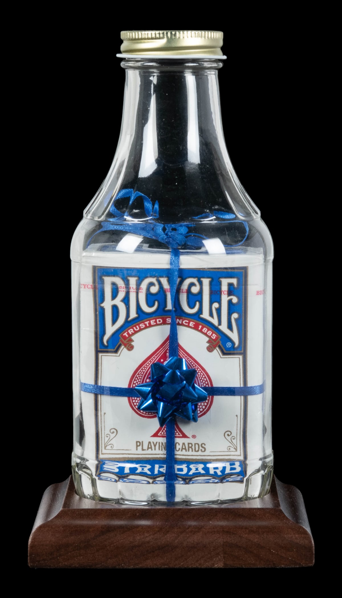 Blue Giftwrapped Deck Impossible Bottle.: Blue Giftwrapped Deck Impossible Bottle. Arizona: Jeff Scanlan, 2020s. Empty glass bottle contains a full sealed pack of card wrapped in a blue mylar ribbon terminating in a bow; the ribbon is