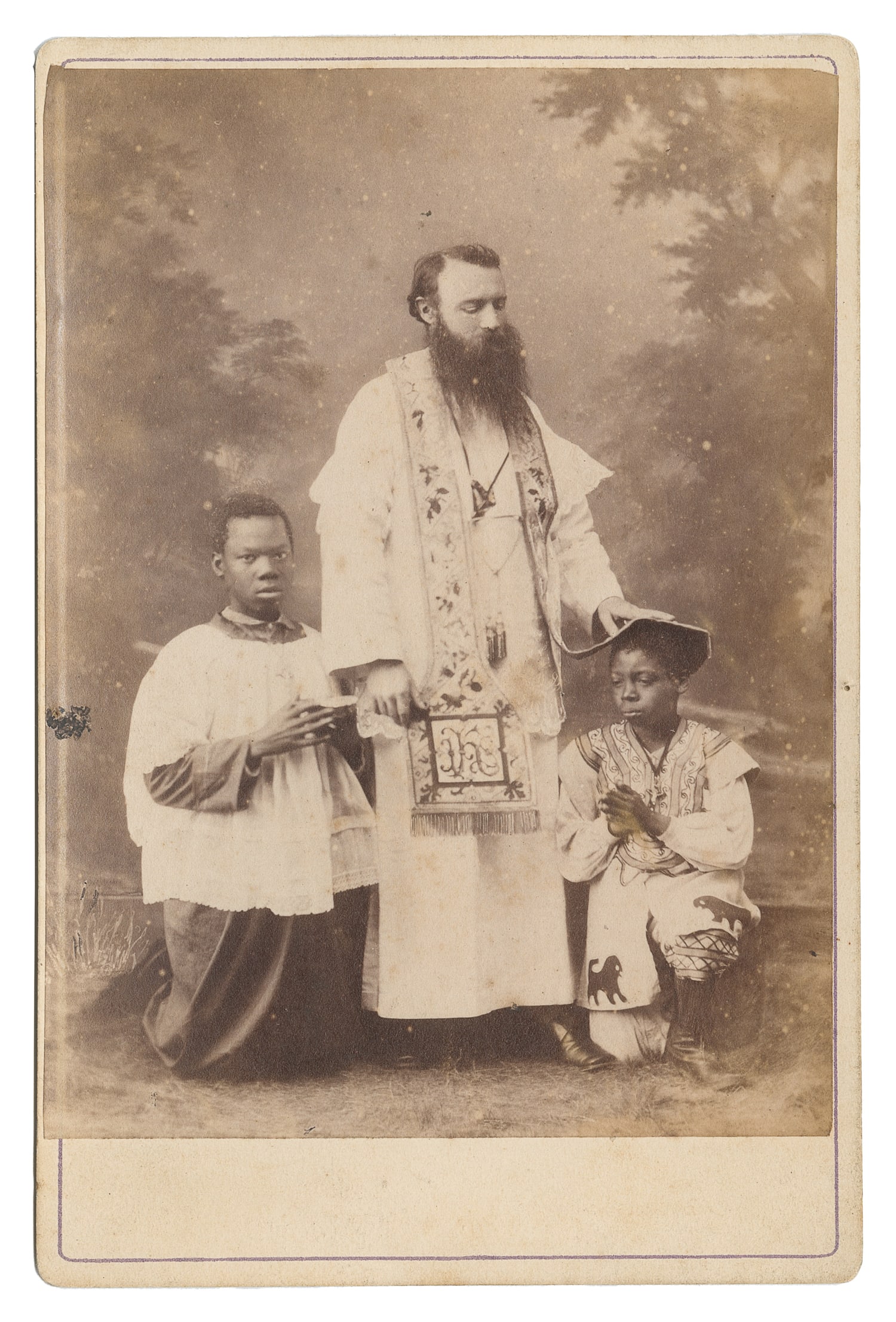 Cabinet photo of a priest and two Black boys.: Cabinet photo of a priest and two Black boys. N.p., ca. 1890s. Portrait of a bearded priest standing, with the boys kneeling at his side, all three in religious clothing. 6 3/8 x 4 1/4".