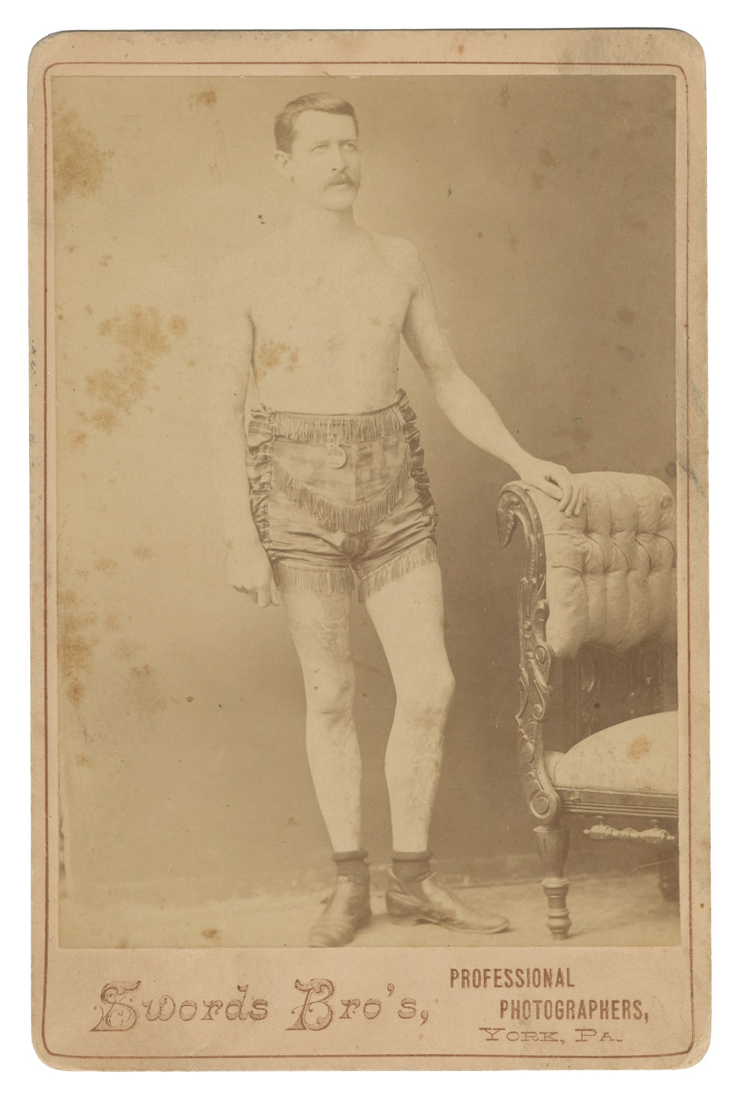 [TATTOO]. Cabinet Card of Tattooed Man. Circa late 19th century.: [TATTOO]. Cabinet Card of Tattooed Man. York, P.A.: Swords Bro's, ca. late 19th century. Albumen print on studio mount. Full-length portrait of a tattooed man wearing fringed shorts. 6 1/ 2 x 4 1/4".