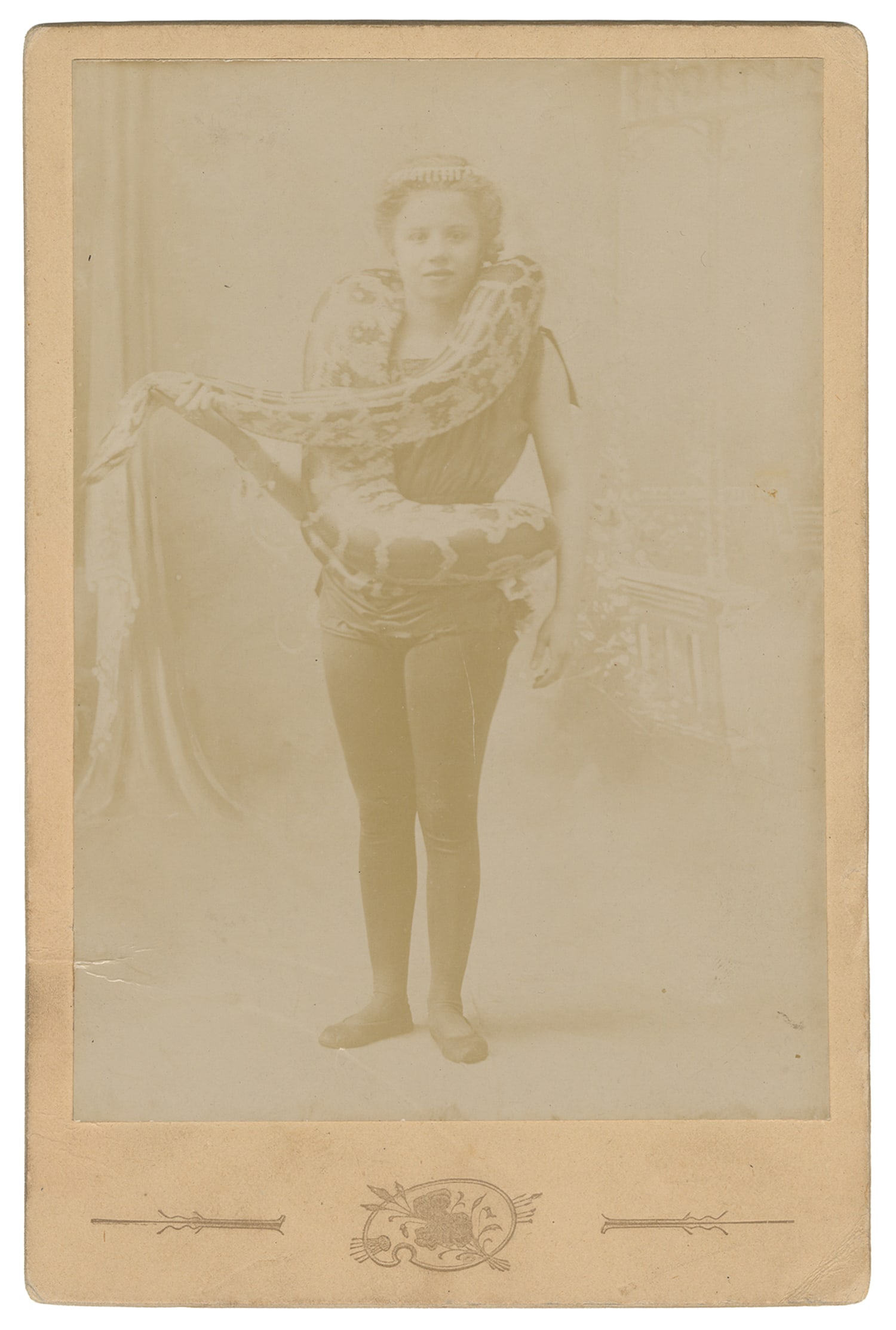 Cabinet photo of a child snake charmer.: Cabinet photo of a child snake charmer. N.p., ca. 1900s. Photograph on card mount, depicting a girl in performing attire with a large snake wrapped around her body. Annotated in pencil on verso: "Mlle
