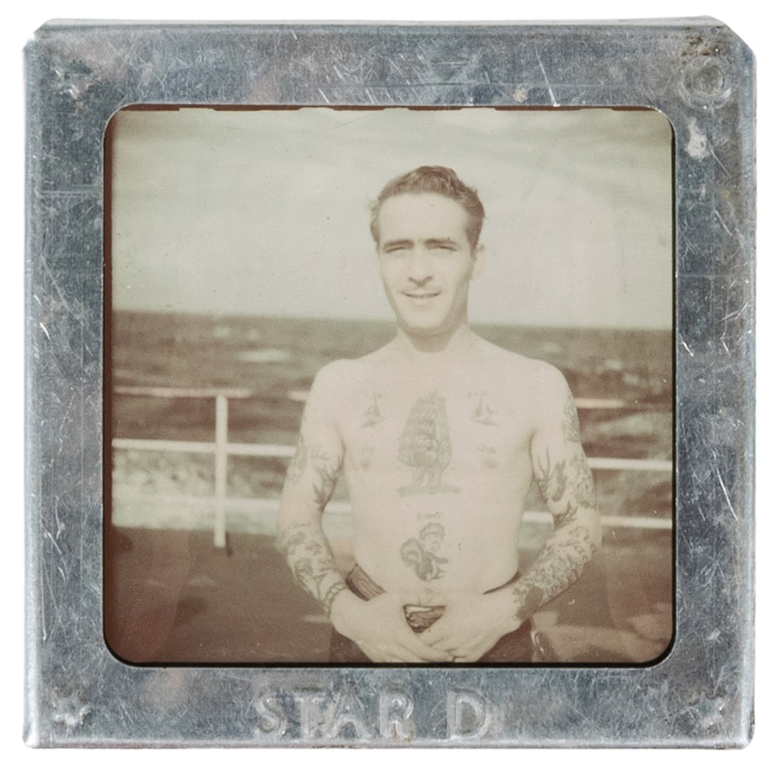[TATTOO]. "Star D" Framed Naval Portrait Negative. U.S., c. 1940s.: [TATTOO]. "Star D" Framed Naval Portrait Negative. U.S., c. 1940s. Miniature tin frame housing a transparency slide of a shirtless gentleman showcasing his tattoos of traditional iconography, includin