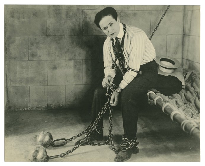 Houdini, Harry. Harry Houdini jail cell escape photo.: Houdini, Harry. Harry Houdini jail cell escape photo. Los Angeles, 1918. Used to publicize Houdini’s serial movie The Master Mystery, he sits in a prison cell wearing cuffs, leg irons, and shackles.
