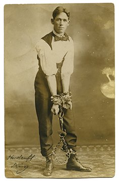 Houdini Imitator. Photo postcard of a "handcuff king": [Houdini Imitator] Real photo postcard of a “handcuff king.” American, ca. 1907. The man stands in a Houdini-esque pose, wrists covered in handcuffs, which are linked to a set of leg irons. Divide