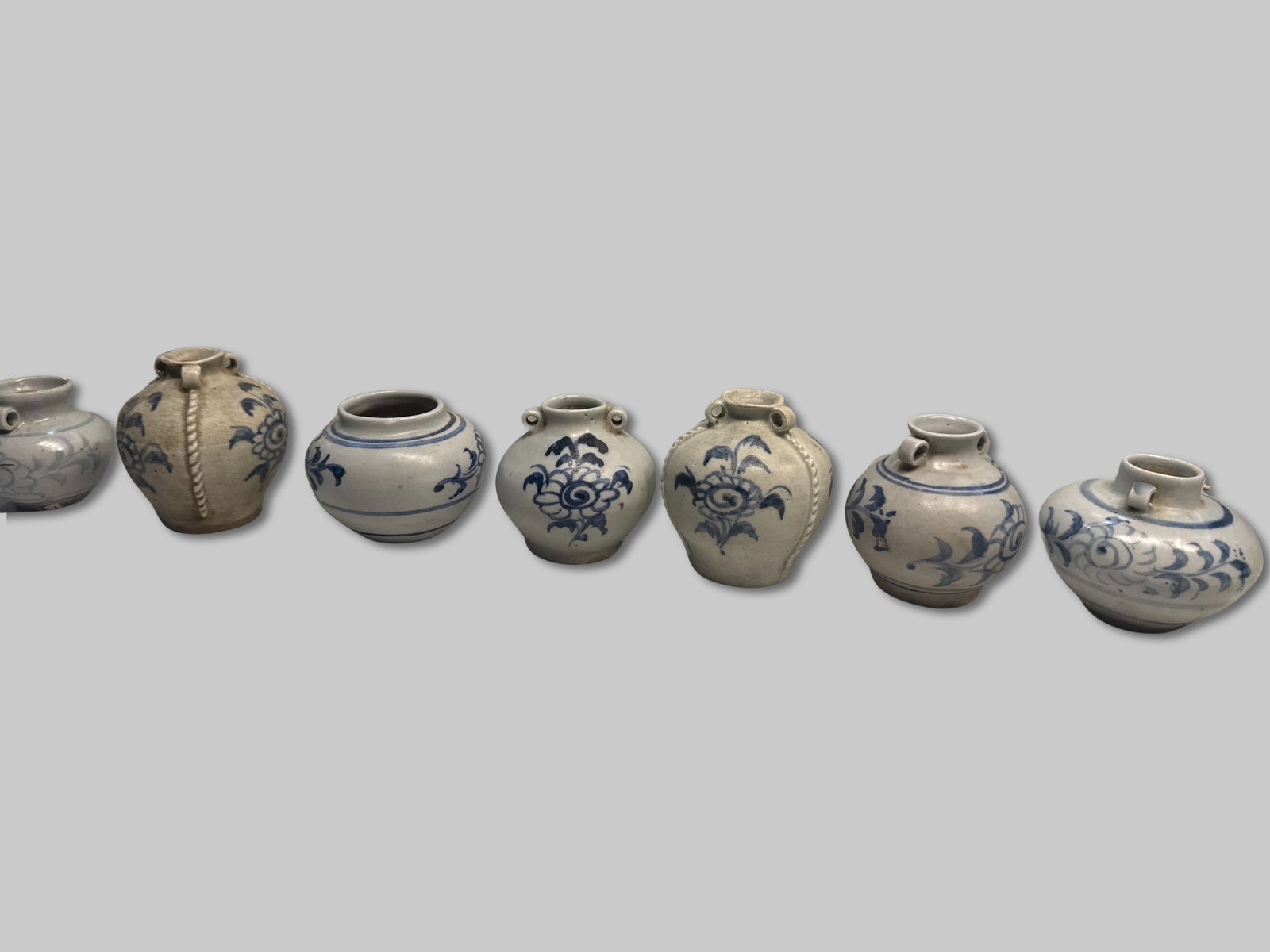 Type Series of Small Blue-and-White Jarlets with Lug Variants Qty 7 -2 1/2" x 3 1/2": 7 small Southeast Asian jarlets featuring hand-painted cobalt blue underglaze floral and scroll motifs. Most are globular with short necks and narrow mouths. Lug configuration varies within the group�