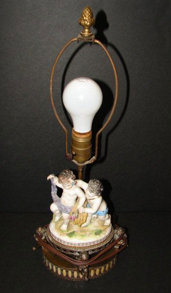 FIGURAL PORCELAIN LAMP: FIGURAL PORCELAIN TABLE LAMP. Figural porcelain lamp depicts a pair of cherubs with wheat attached to a brass base with three attached cherubs. Marked with blue mark on reverse of porcelain. Lamp is e