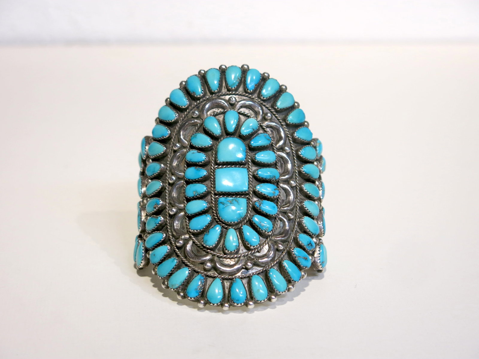 J&E Wilson Navajo Sleeping Beauty Turquoise Cuff: A classic Navajo rosette/sunburst layout of radiating teardrop stones around a central squared cushion cabochon, all within a stamped and repoussé silver ground with scroll and floral elements. The s
