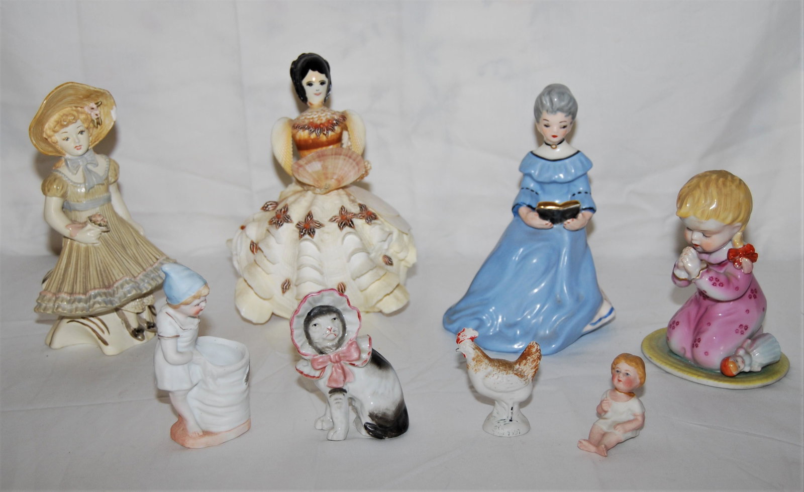 Vintage Bisque, Porcelain & Seashell Figurines: Vintage Bisque & Porcelain Figurines & a Unique Sea-Shell Figure. 9 figures are in this lot including: a seated woman in a blue dress reading, 7" tall, also - a young girl kneeling and praying, 5" tal