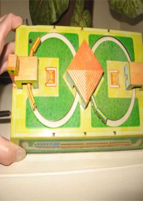 Wind up Toy Train: Tin wind up train toy, w/ key, aprox. 9 1/2 w x 4 5/8 w x 2 5/8 tall, 2 trains on circular tracks w/ 3 buildings, runs, makers mark appears to be a V within a circle