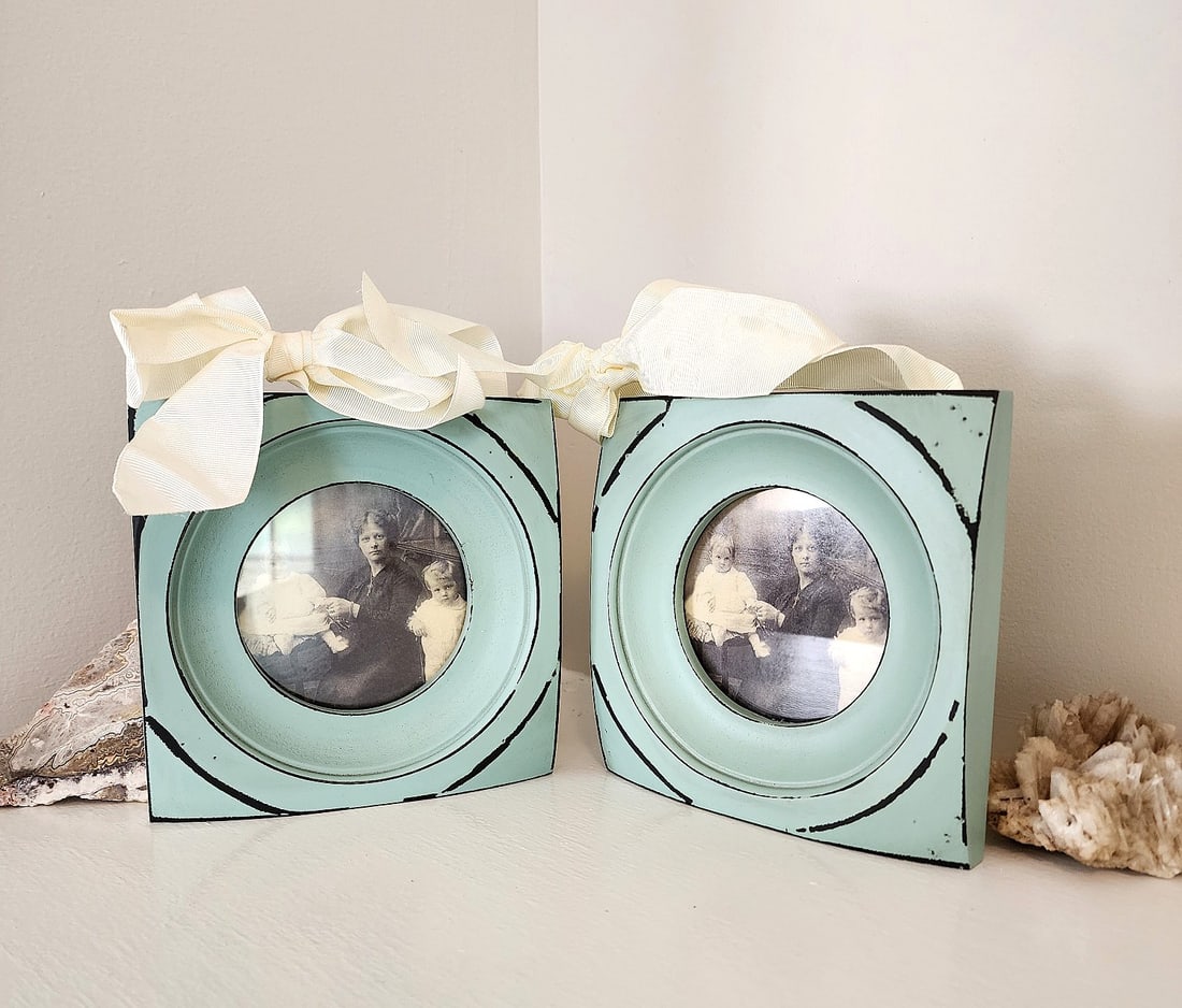 2 Light Blue Distressed Picture Frames: 2 New faux distressed picture frames. Square. Round display with glass. Hinge flap back opening. Features wide, grosgrain ivory ribbon bows for hanging. Measures: 6 1/4 x 6 1/4.