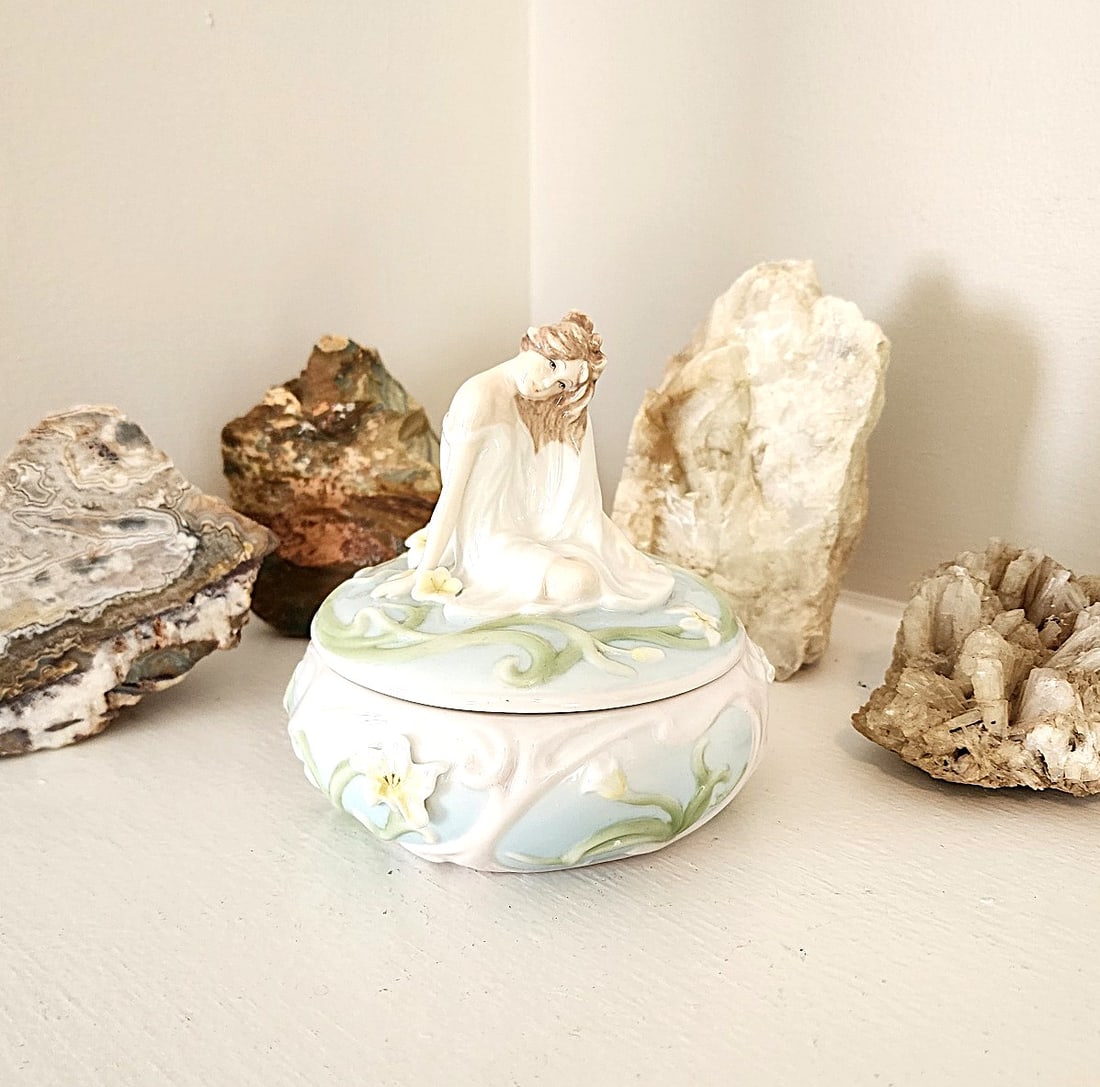 Fine Porcelain Trinket/jewelry Box with girl sitting on top.: Pastel colors. The base features lilies and butterflies. New. From Unicorn Studios. Measures: approx. 5" x 5".