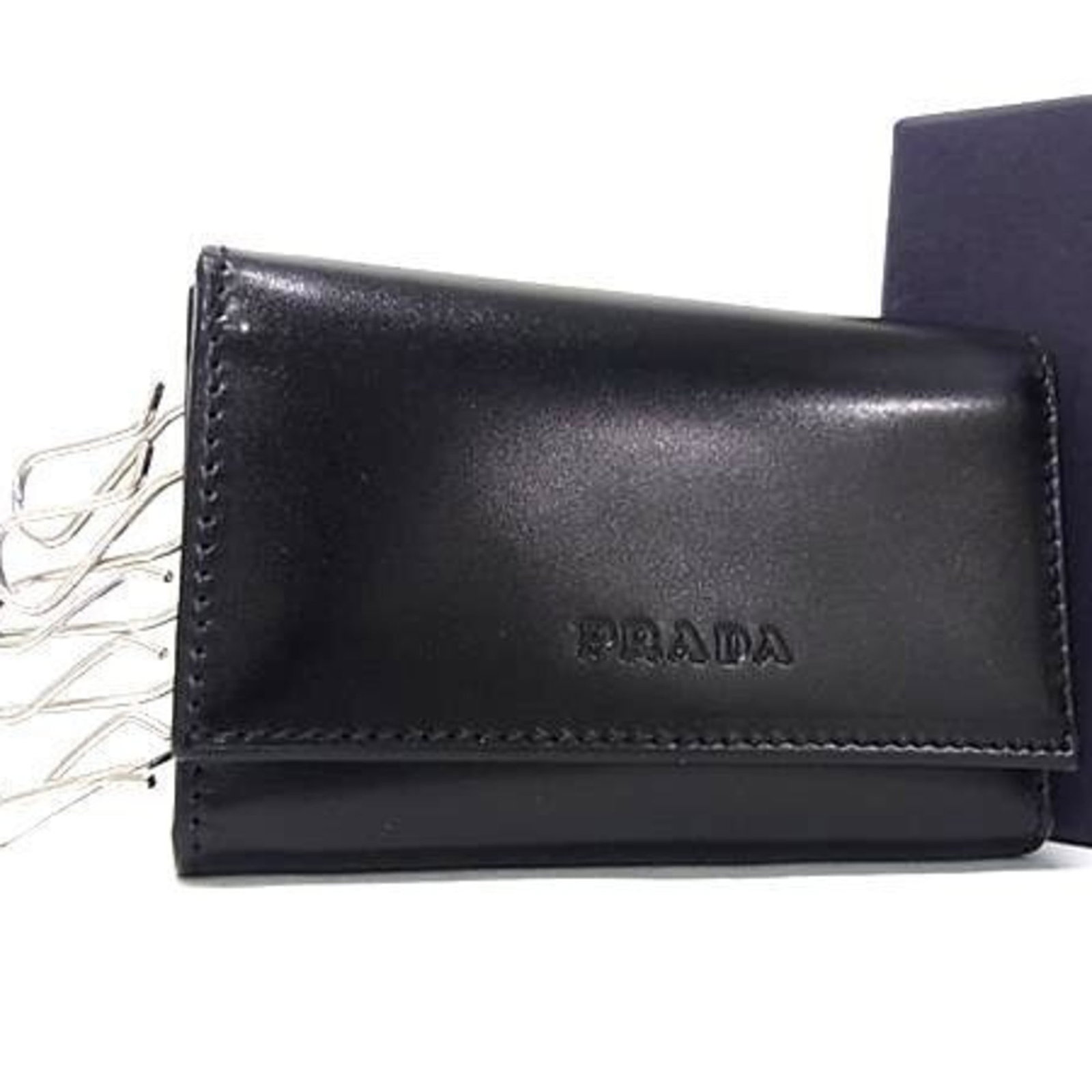Prada Keycase Leather: Prada Keycase Leather Black Prada Keycase Leather in very good condition, with some scratches and dirt visible. A few traces of usage are present, adding to its worn-in feel. Overall, this keycase exu