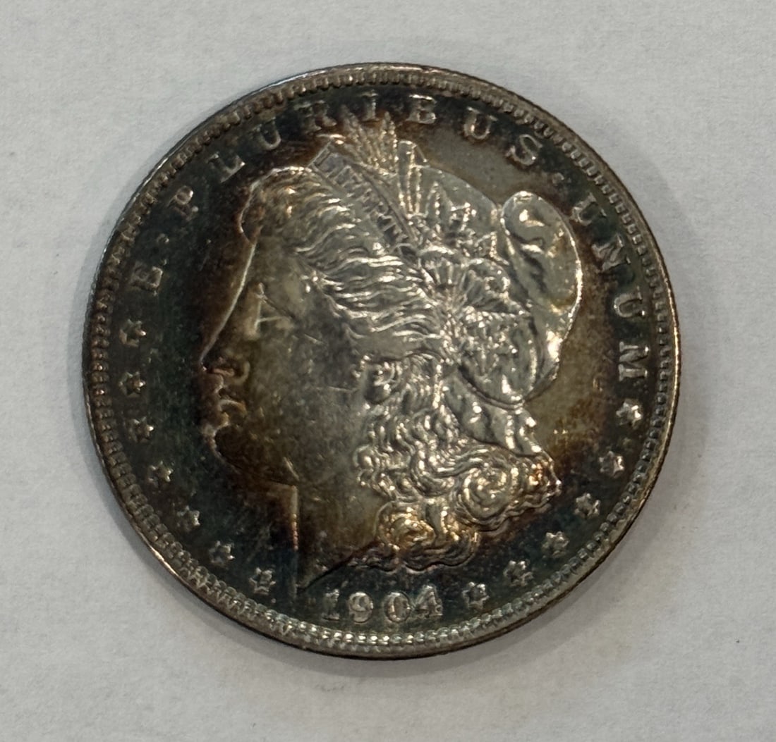 MORGAN DOLLAR CHOICE AU 1904 RAINBOW TONING: MORGAN DOLLAR CHOICE AU 1904 RAINBOW TONING The 1904 Rainbow Toning Morgan Dollar Choice AU features a unique display of toning on its surface, adding visual interest to this classic silver coin. With