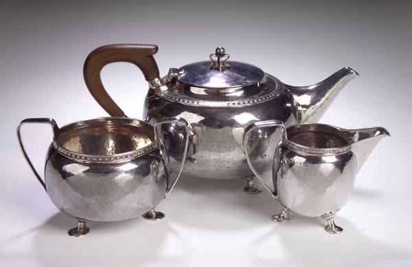An A.E. Jones three piece silver tea set: An A.E. Jones Arts and Crafts three piece silver tea service, comprising a tea pot; a milk jug; and a sugar bowl, each of ovoid form with rosette band and hammered ground raised above three pad feet,