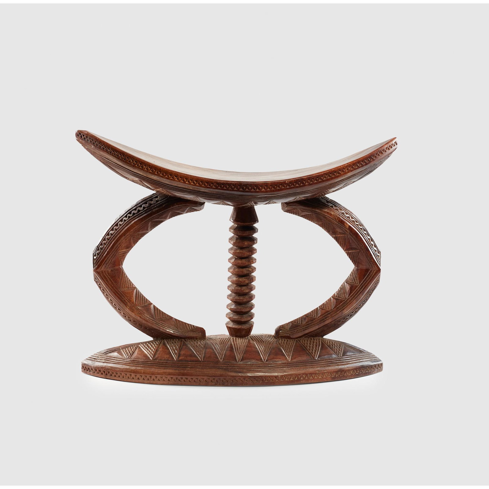 IGBO STOOL NIGERIA: carved wood, standing on a elliptical base, with central ribbed support and dual outer supports, the seat slightly curved and with tapering edges, carved detailing throughout (Dimensions: 40cm tall) (