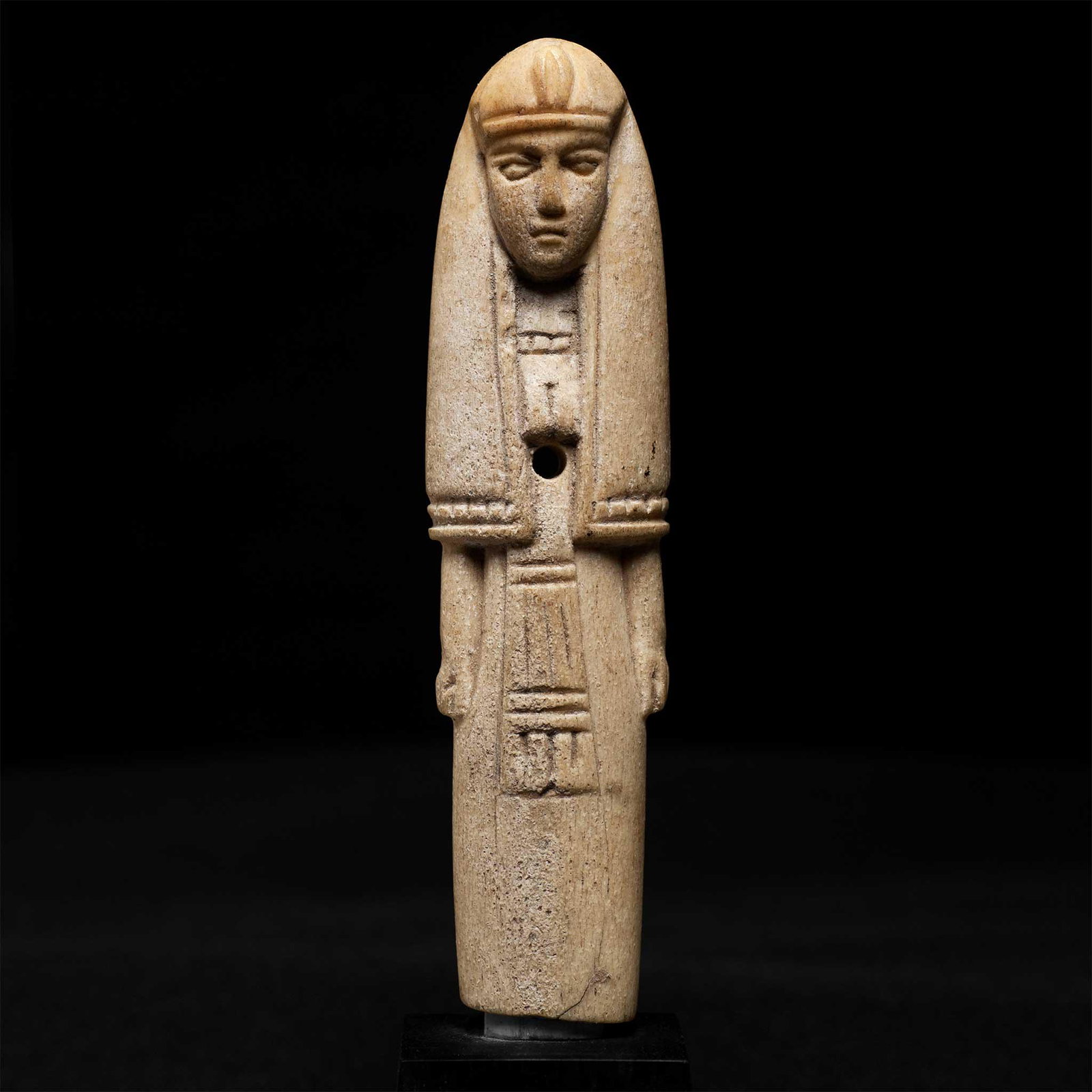 ANCIENT EGYPTIAN BONE FIGURE EGYPT, ROMAN PERIOD, C. 1ST CENTURY A.D.: carved bone, the standing figure wears a long wig and belted dress, perforated in the centre for attachment, raised on bespoke mount (11.5cm tall) Ex. collection Jeanne et Pierre Vérité, Paris (1930