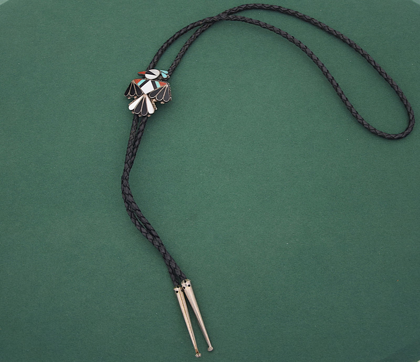 NATIVE AMERICAN STYLE INLAY THUNDERBIRD BOLO TIE, SILVER: DECORATIVE BOLO TIE FEATURING A STRIKING THUNDERBIRD MOTIF WITH INLAID STONE OR SHELL IN BLACK, WHITE, TURQUOISE, AND RED TONES, SET IN A SILVER MOUNT; THE SLIDE IS ATTACHED TO A BLACK BRAIDED CORD WI