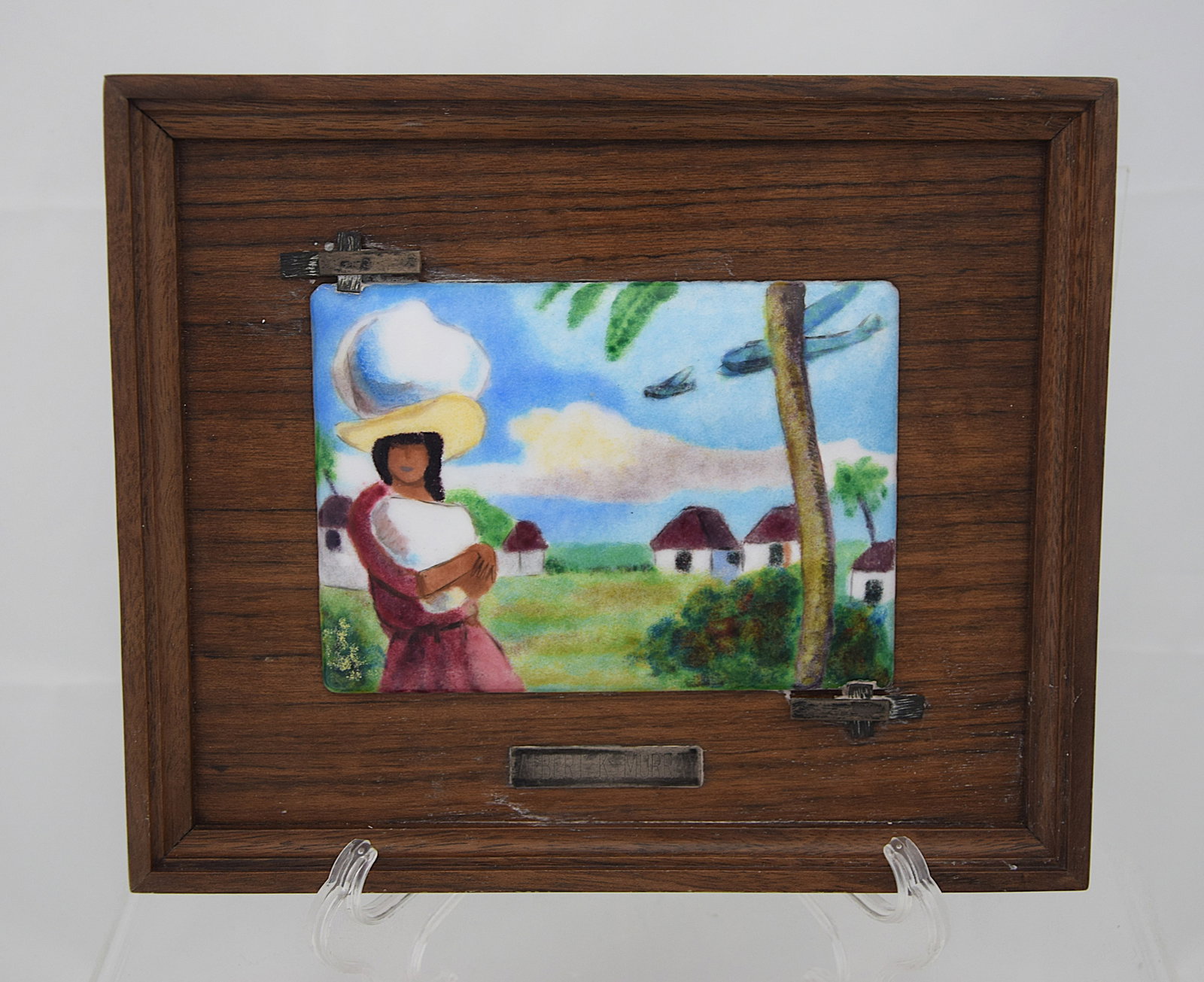 FINE ENAMEL PLAQUE CARIBBEAN FIGURAL SCENE AFTER ALBERT K. MURRAY FRAMED: VIBRANT HAND-PAINTED ENAMEL PLAQUE DEPICTING A WOMAN IN A TROPICAL LANDSCAPE BALANCING A VESSEL, WITH VILLAGE HOUSES AND LUSH GREENERY IN THE BACKGROUND. AFTER THE STYLE OF ALBERT K. MURRAY, PRESENTED