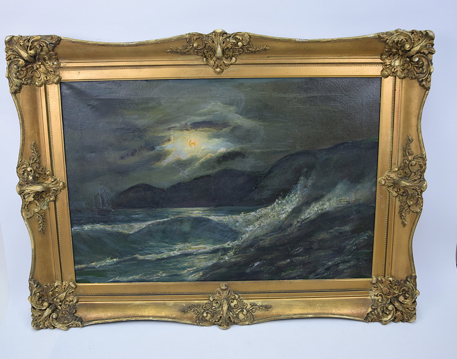 ANTIQUE OIL PAINTING MOONLIT SEASCAPE IN ORNATE GILT FRAME POSSIBLY 19TH–EARLY 20TH CENTURY: ATMOSPHERIC ANTIQUE OIL ON CANVAS DEPICTING A DRAMATIC MOONLIT SEASCAPE, WITH ROLLING WAVES CRASHING IN THE FOREGROUND AND A SMALL SAILBOAT VISIBLE IN THE DISTANCE UNDER A GLOWING BREAK IN THE CLOUDS;