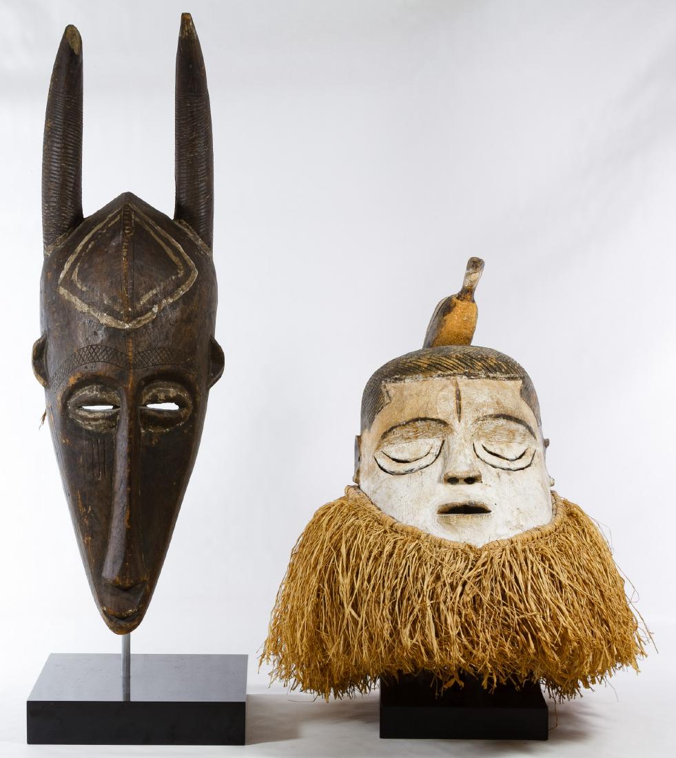 African Carved Wood Masks: African Carved Wood Masks Two items including a head with carved bird on top and white painted face with grass collar and an oblong horned mask with diamond painted forehead; both have display stands 
