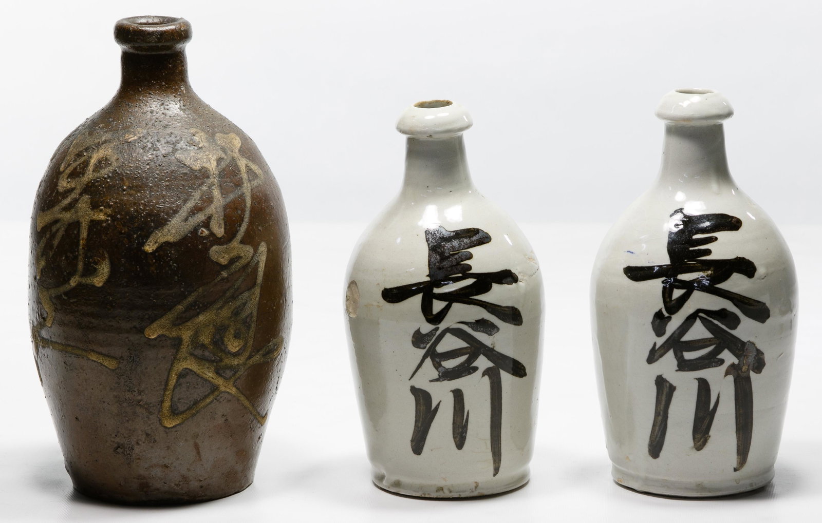 Japanese Tokkuri Sake Bottle Assortment: Japanese Tokkuri Sake Bottle Assortment Three early 20th century items including a brown Tamba ware bottle having white slip-trail kanji decoration and two white glazed Tamba kiln bottles having black