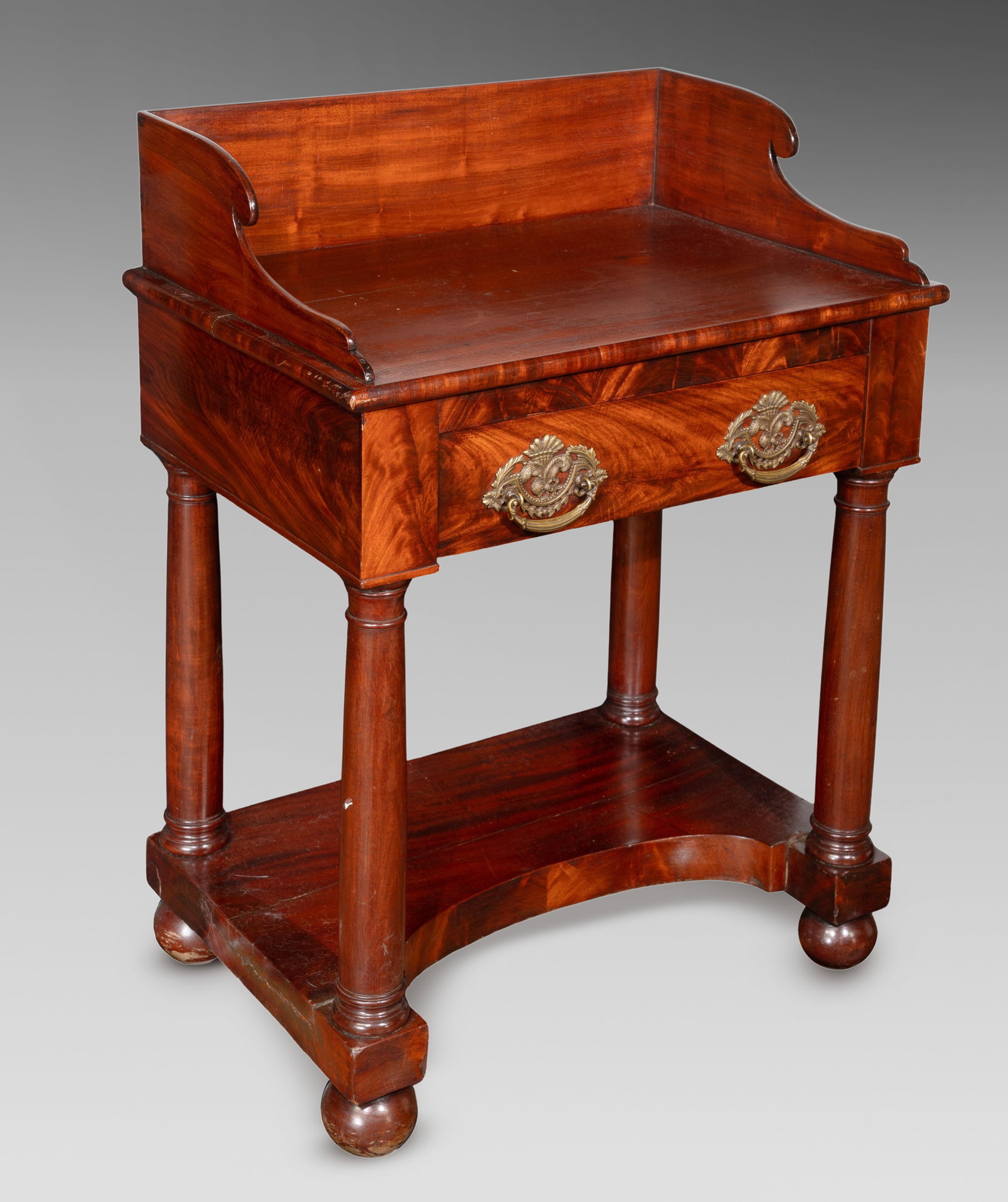 American Empire Mahogany Wash Stand: American Empire Mahogany Wash Stand Having a curvilinear back splash, a dovetailed drawer having cast brass pulls, supported by columnar legs above a shelf / stretcher, on bun feet Property from: an