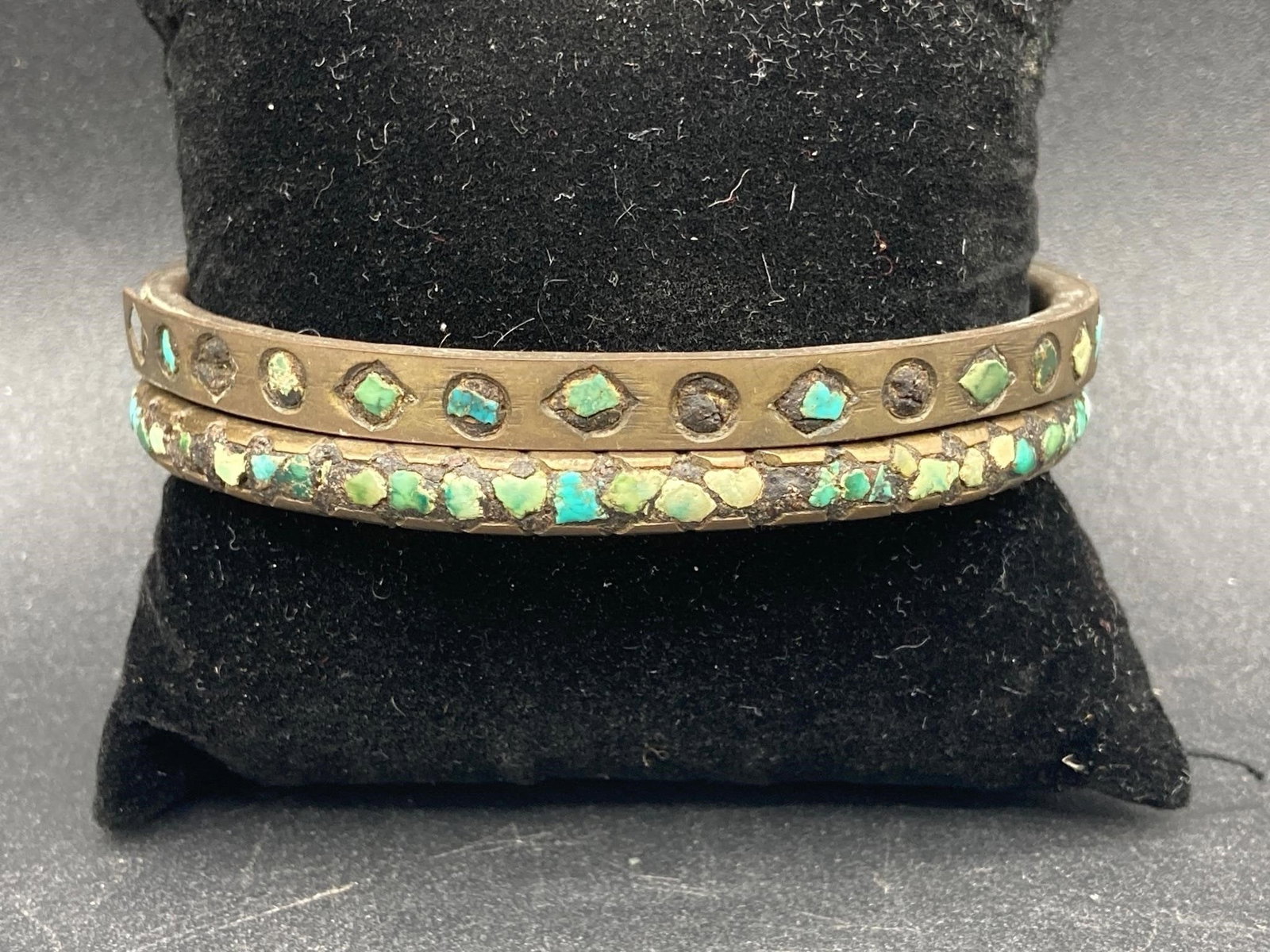 Turquoise Stone Brass Bangles: Pair of brass bangles. Bangles feature multiple inlayed stone fragments including turquoise stones. Both measure 3" wide. Some areas of missing stones and one bracelet has a small area of separation a