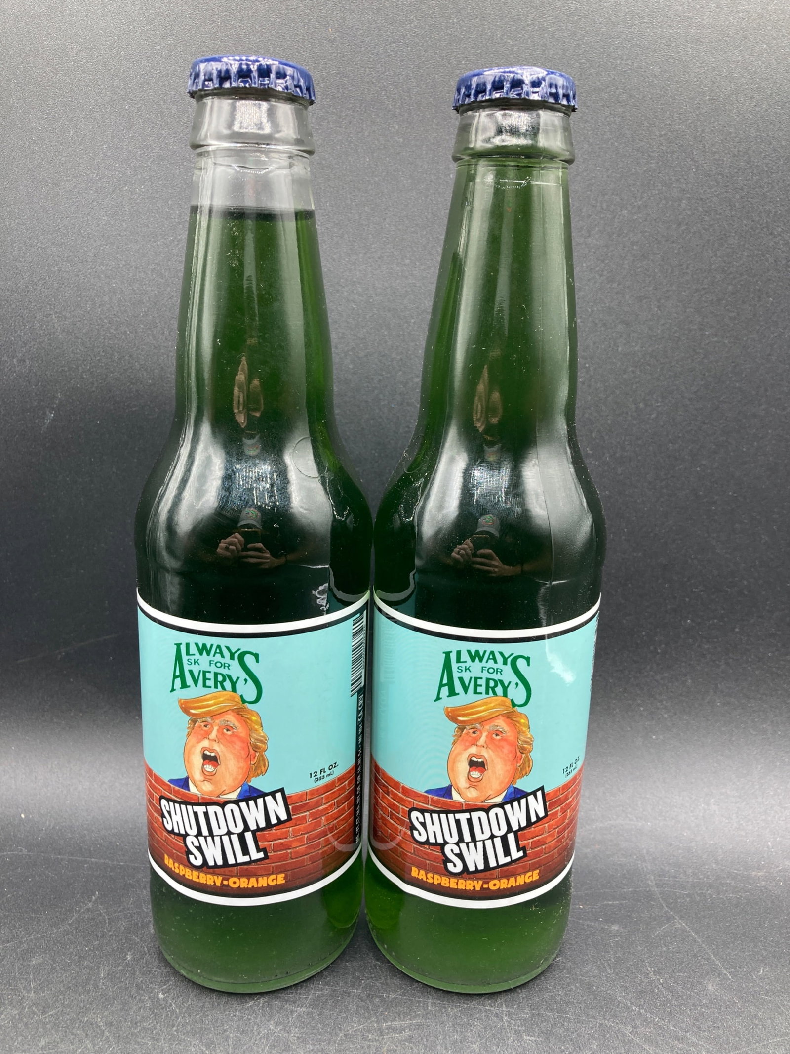 Set of 2 Avery's Shutdown Swill Donald Trump Sodas: This listing features a set of two unopened Avery's 'Shutdown Swill' raspberry-orange flavored sodas, each contained in a 12 fl oz glass bottle. These bottles are products from Avery's Beverages, a co