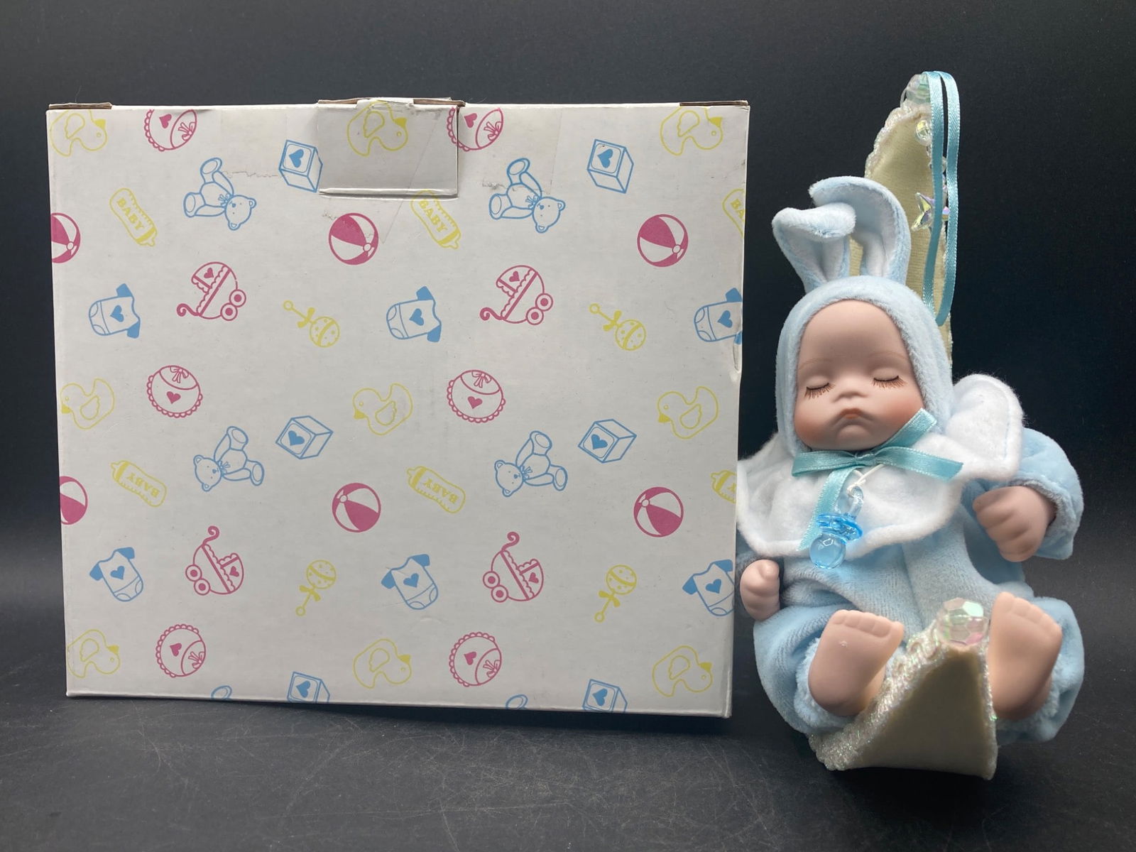 Baby Doll Music Box On Moon-Shaped Swing: This listing features a baby doll music box seated on a moon-shaped swing with its original box. The baby doll is dressed in a light blue outfit with detailing that includes a pacifier accessory and r