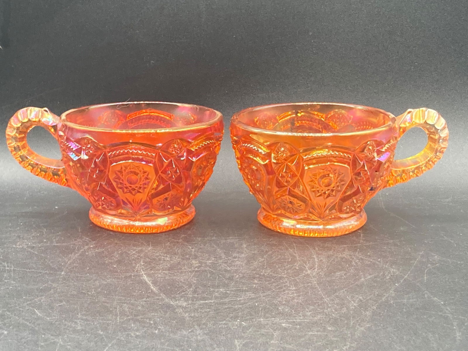 Carnival Glass Marigold Heirloom Tea Cups: This listing features a pair of vintage carnival glass tea cups in the marigold color. These tea cups are designed with the heirloom pattern, showcasing intricate floral and geometric motifs, characte