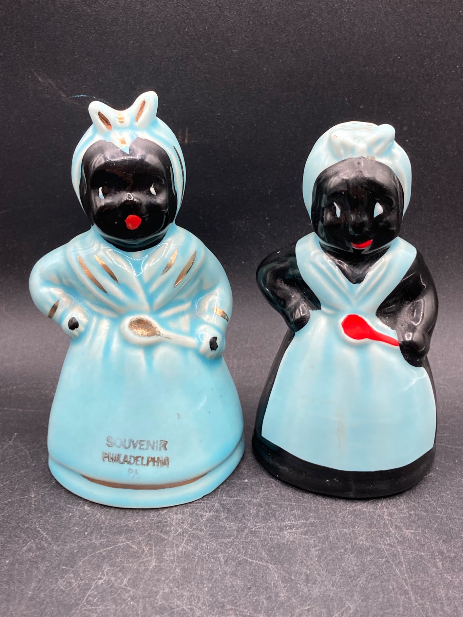 Black Americana Porcelain Salt & Pepper Shakers: Two porcelain salt and pepper shakers featuring Black Americana imagery, with both figures dressed in blue and black. One shaker has œSouvenir Philadelphia PA inscribed at the bottom front.