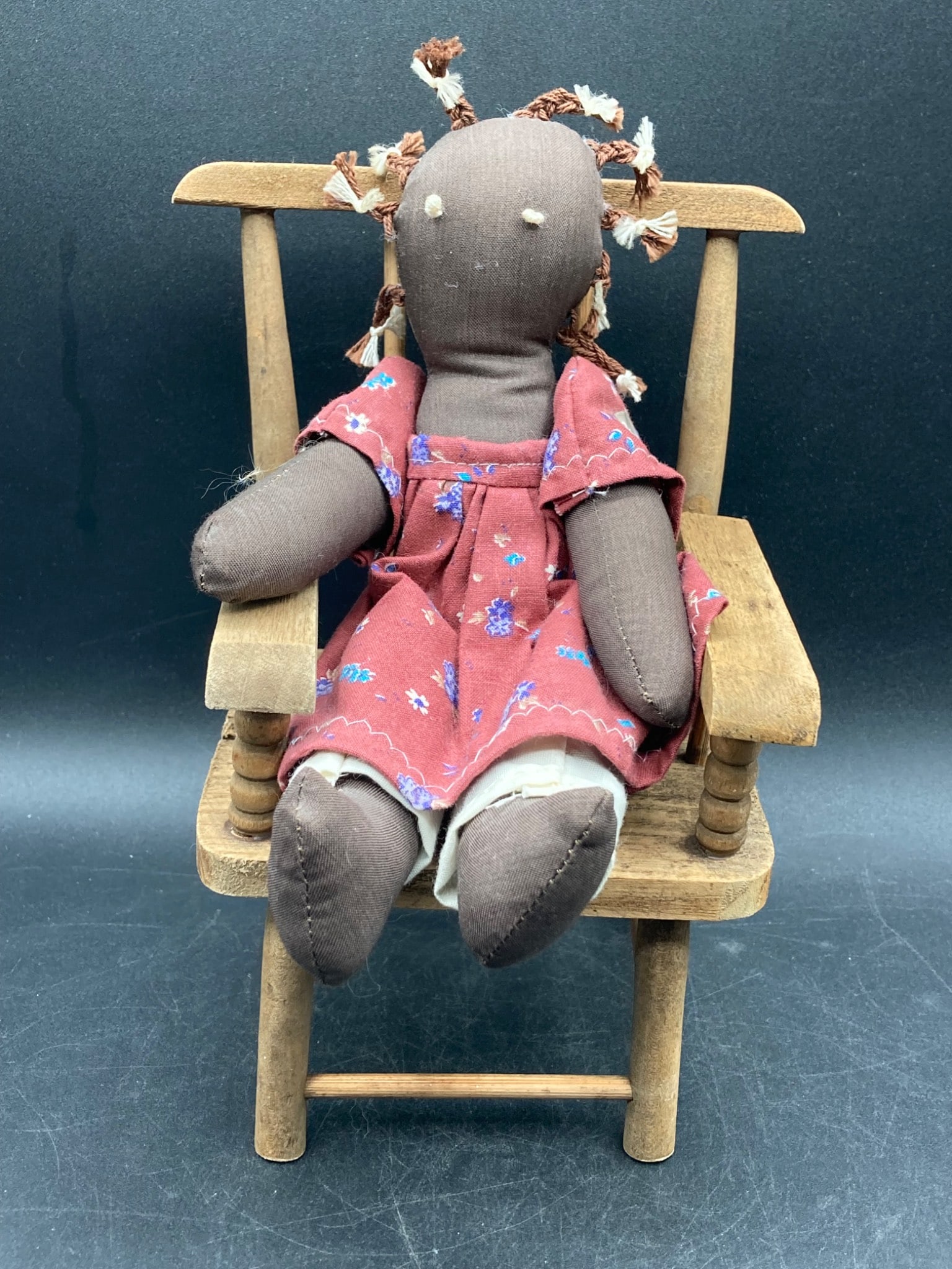 Black Americana Cloth Doll & Wooden Chair Set: This lot includes a handmade Black Americana cloth doll and a wooden chair. The doll features braided yarn hair, embroidered facial details, and a floral red dress with white undergarments. The chair 