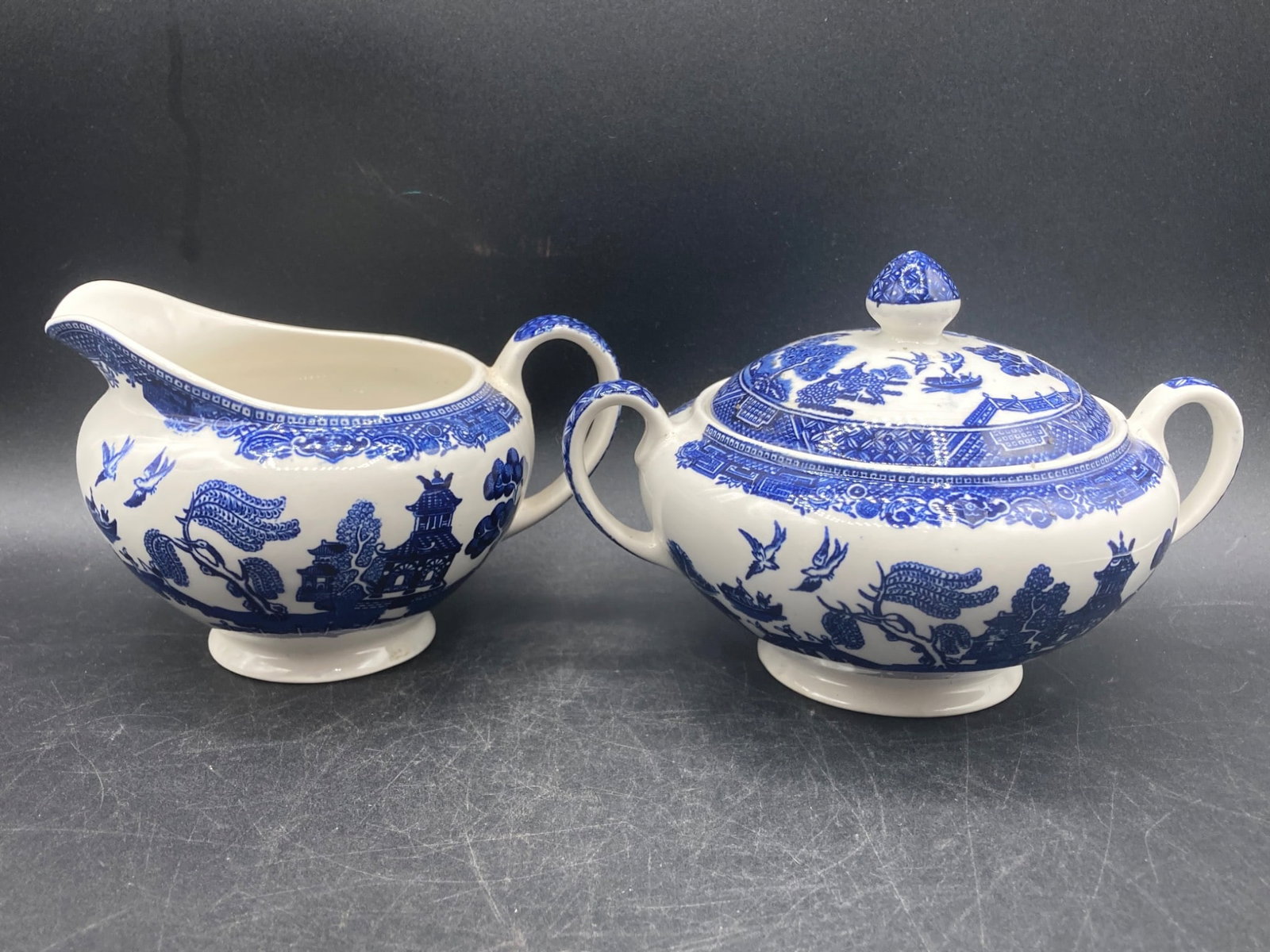 English Porcelain Blue Willow Creamer and Sugar Bowl Set: An English porcelain creamer and sugar bowl set featuring the Blue Willow pattern in cobalt blue on a white background. The sugar bowl includes a fitted lid and dual handles, while the creamer feature