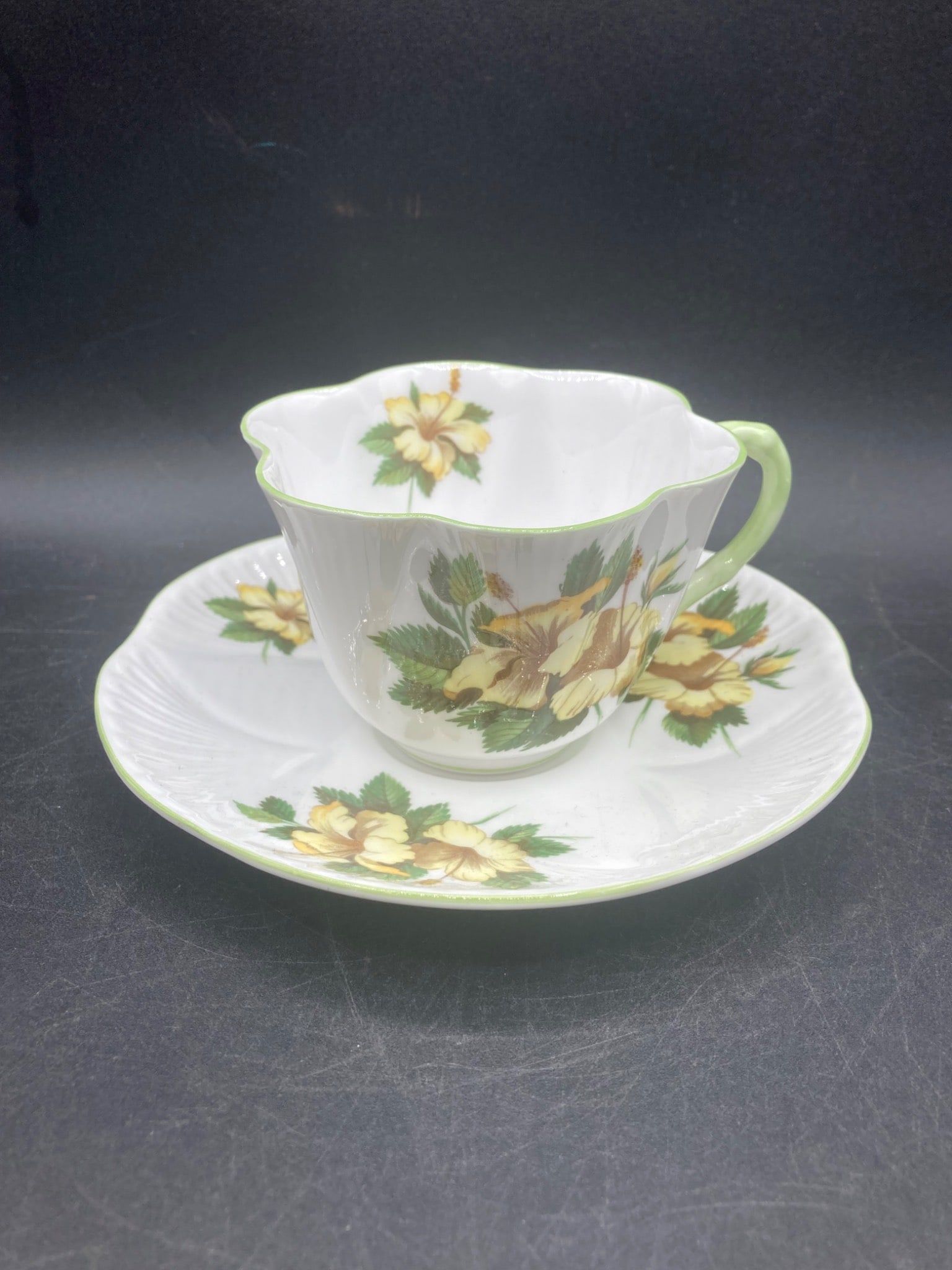 Shelley England Fine Bone China Hibiscus Tea Cup & Saucer: This is a Shelley England Fine Bone China tea cup and saucer in the Hibiscus pattern, number 13882. The set features yellow hibiscus flowers with green foliage, and both the cup and saucer are trimmed