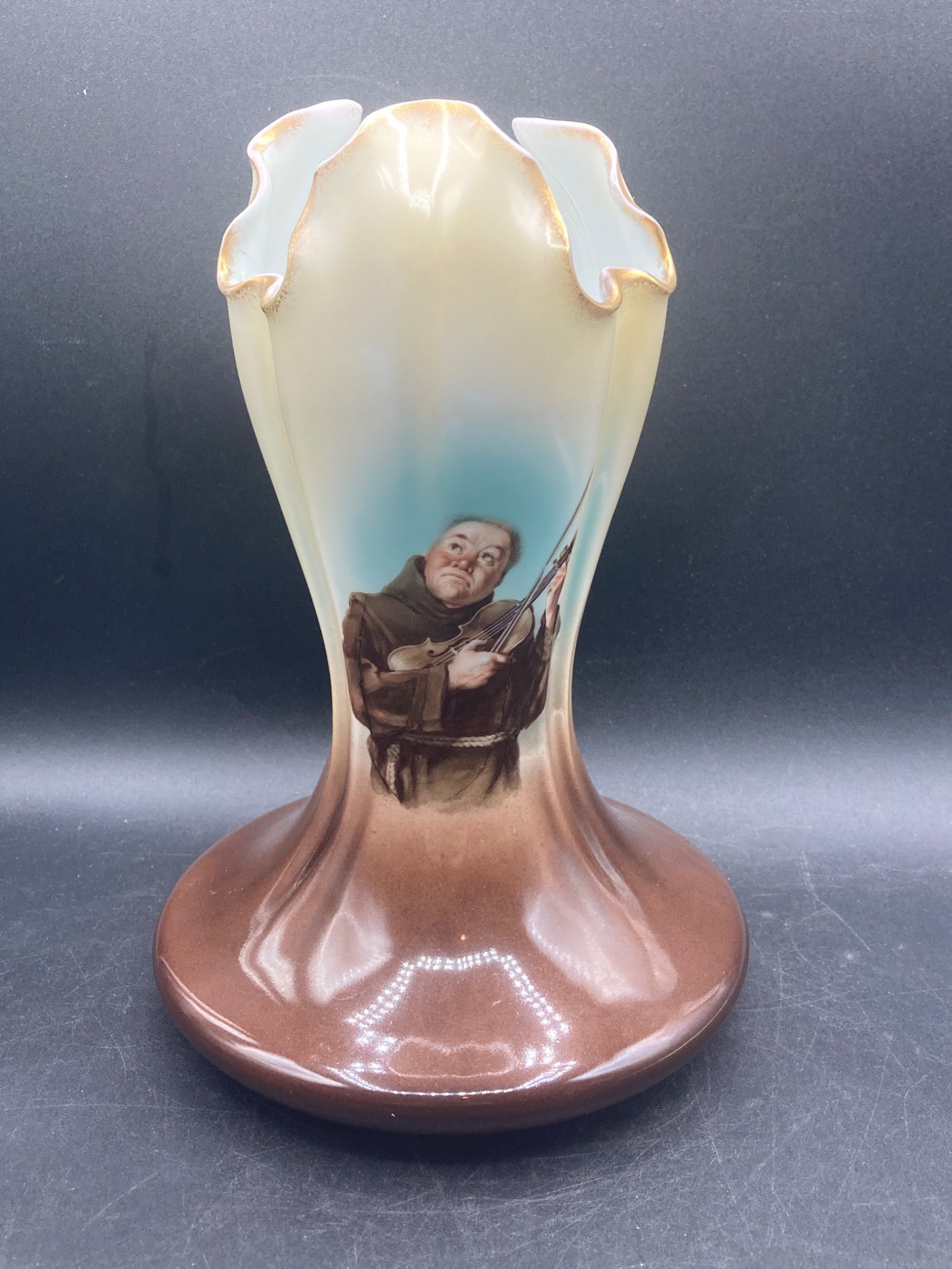 Bavarian Monk Porcelain Vase: This porcelain vase features a depiction of a monk playing a violin, rendered in detailed hand-painted design. The vase has a ruffled rim with gilded edges and a gradient color scheme transitioning fr