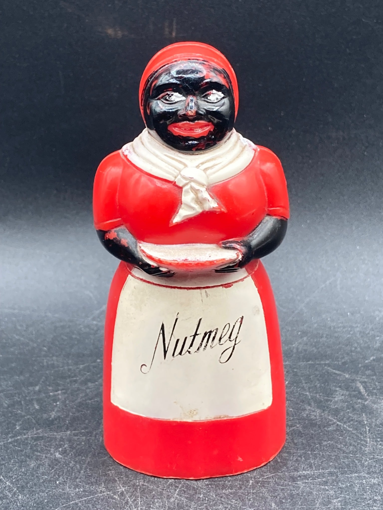 Black Americana Bakelite Nutmeg Shaker: This is a mid-20th century Black Americana nutmeg shaker crafted from Bakelite. It features a red and black painted figure design with 'Nutmeg' written in cursive on the front of the apron. The shaker