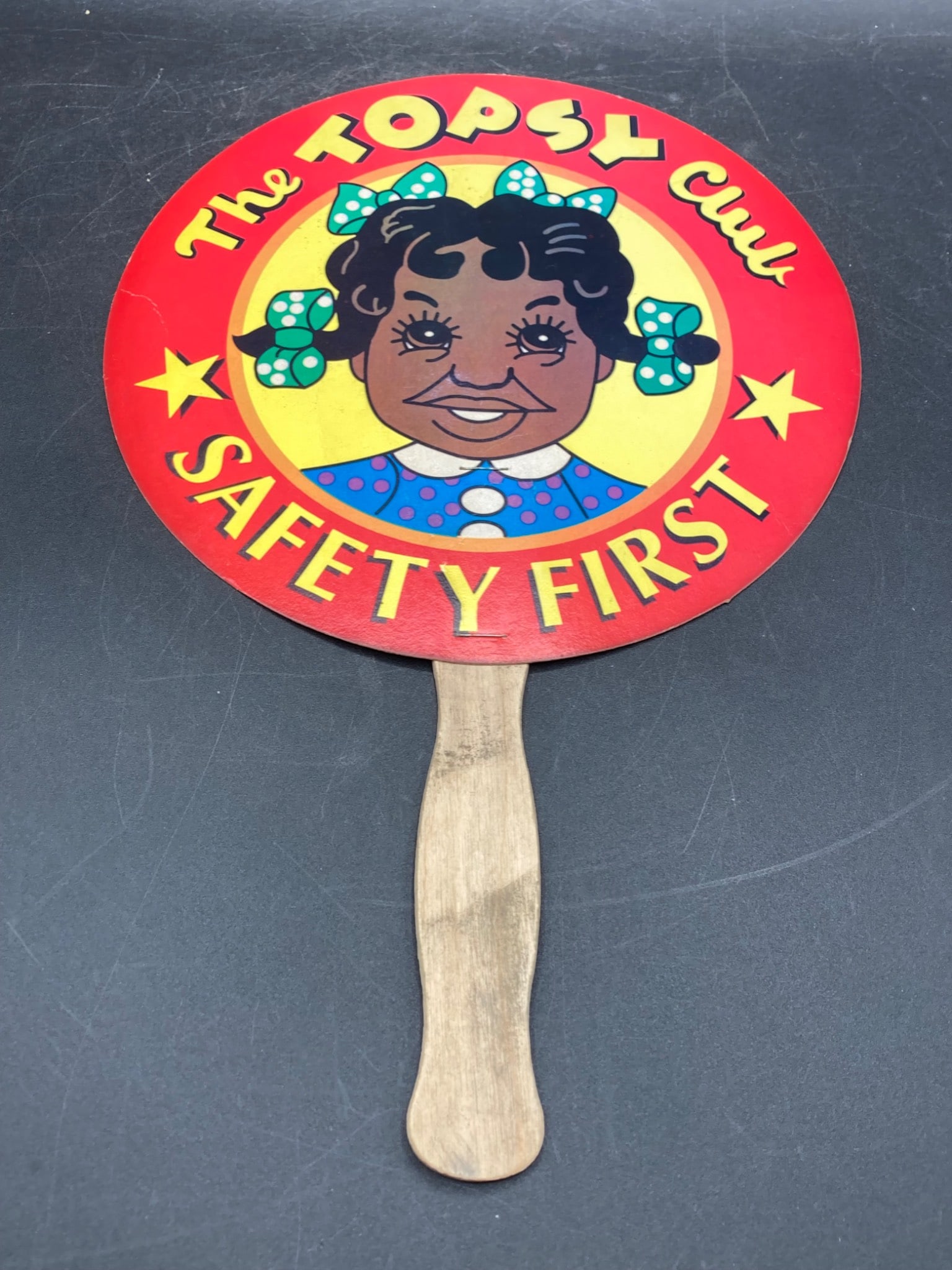 Vintage Black Americana The Topsy Club Fan: This is a promotional fan associated with the Topsy Club, promoting safety awareness. It is constructed with a printed cardboard circular face and a wooden handle. The text "Safety First" and an image