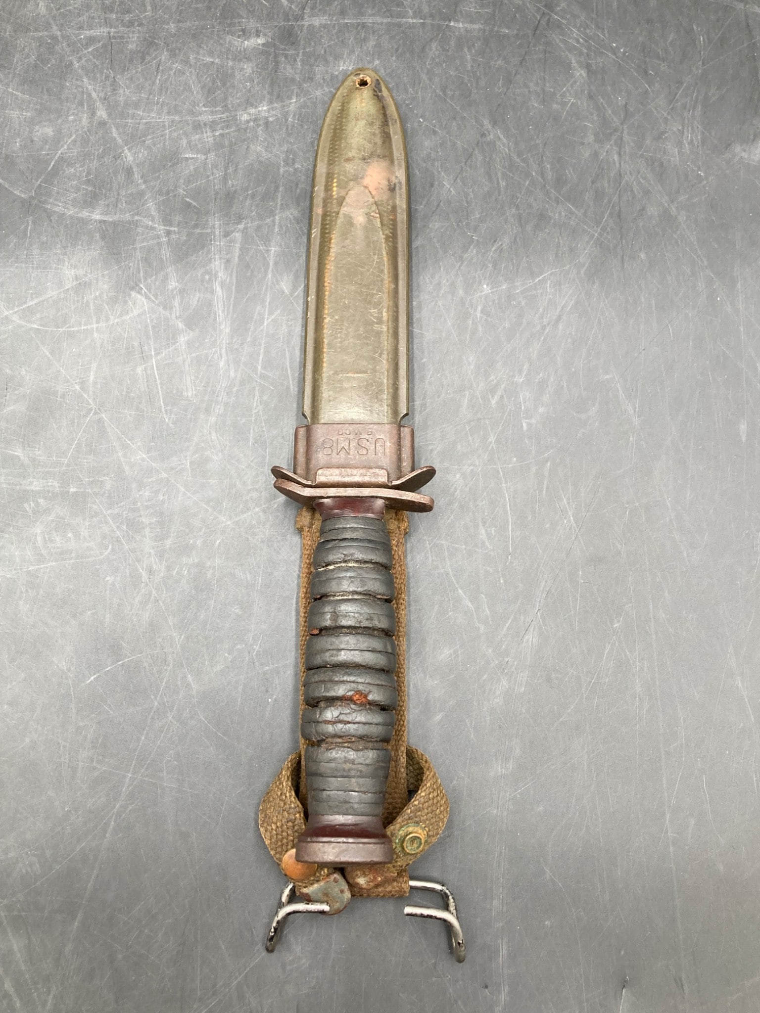 WWII United States M8 Fighting Knife with Sheath and Belt Loop: This is a WWII-era United States M8 fighting knife. The knife features a 6.75-inch steel blade with signs of wear and age-related discoloration. The black, ribbed handle exhibits visible aging with ru