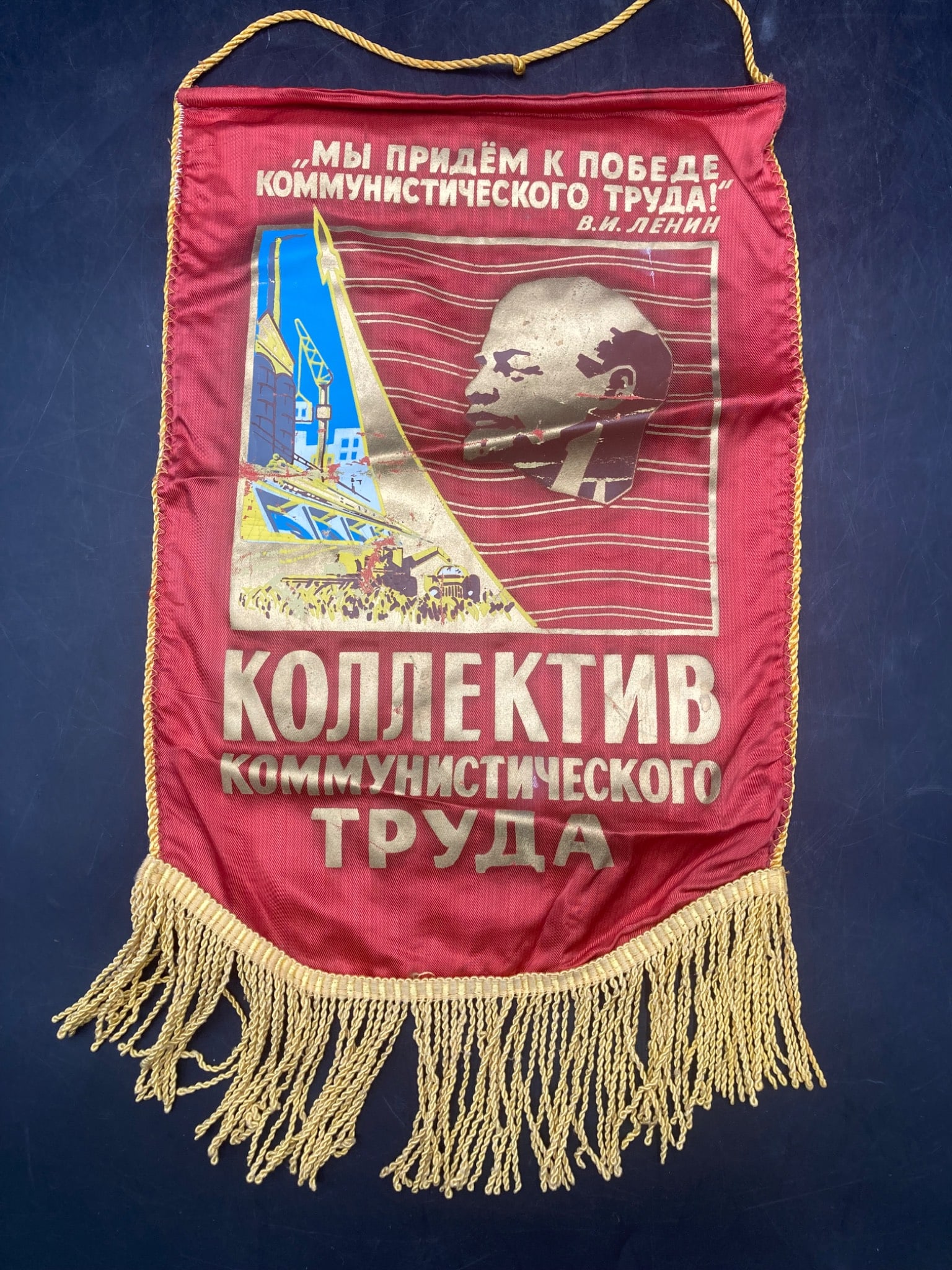 Soviet Union Vladimir Lenin Banner Flag: This is a Soviet-era banner featuring an image of Vladimir Lenin and Russian text promoting collective labor ideals. The banner is made of red fabric with yellow fringe at the bottom. The text transla