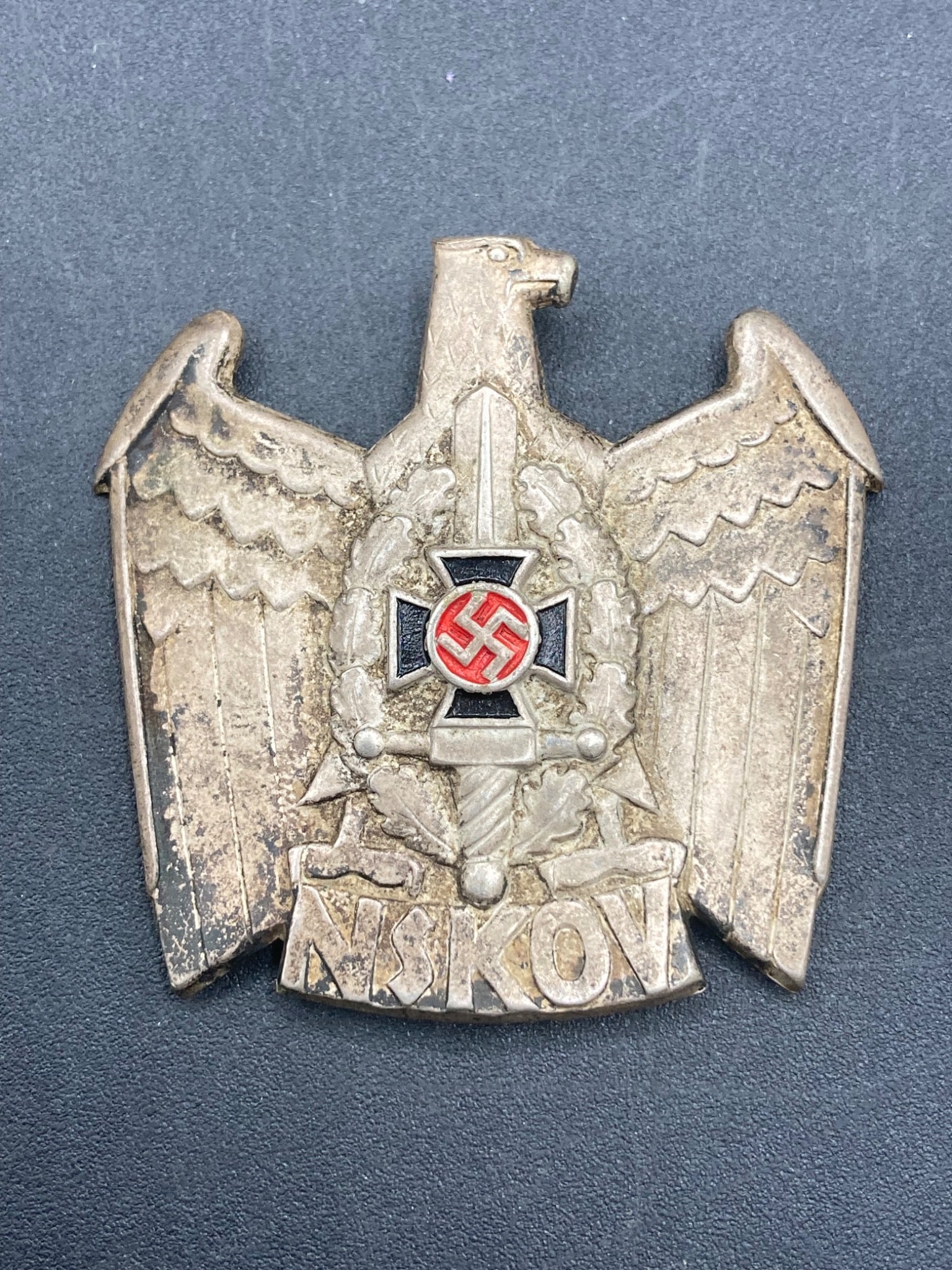 WWII German NSKOV Cap Badge: This lot features an authentic WWII German NSKOV (National Socialist War Victim's Care) cap badge. The badge is constructed of metal with a detailed eagle design holding an Iron Cross containing a swa