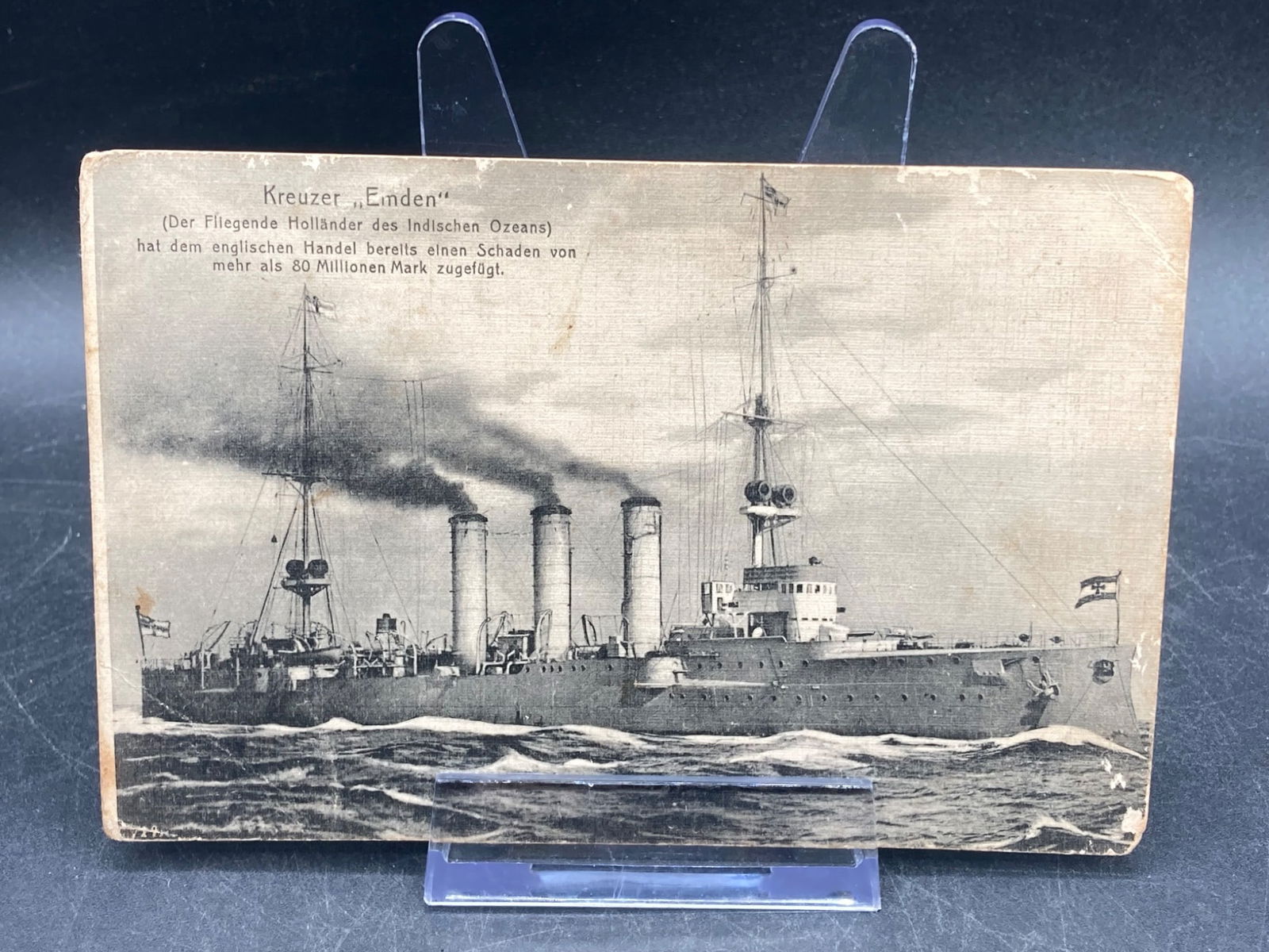1915 German Kreuzer Emden Red Cross Liebesgaben Postcard: A 1915 German 'Kreuzer Emden' postcard, featuring a depiction of the German cruiser SMS Emden. The card includes German text describing the ship's operations and contributions during WWI. The reverse